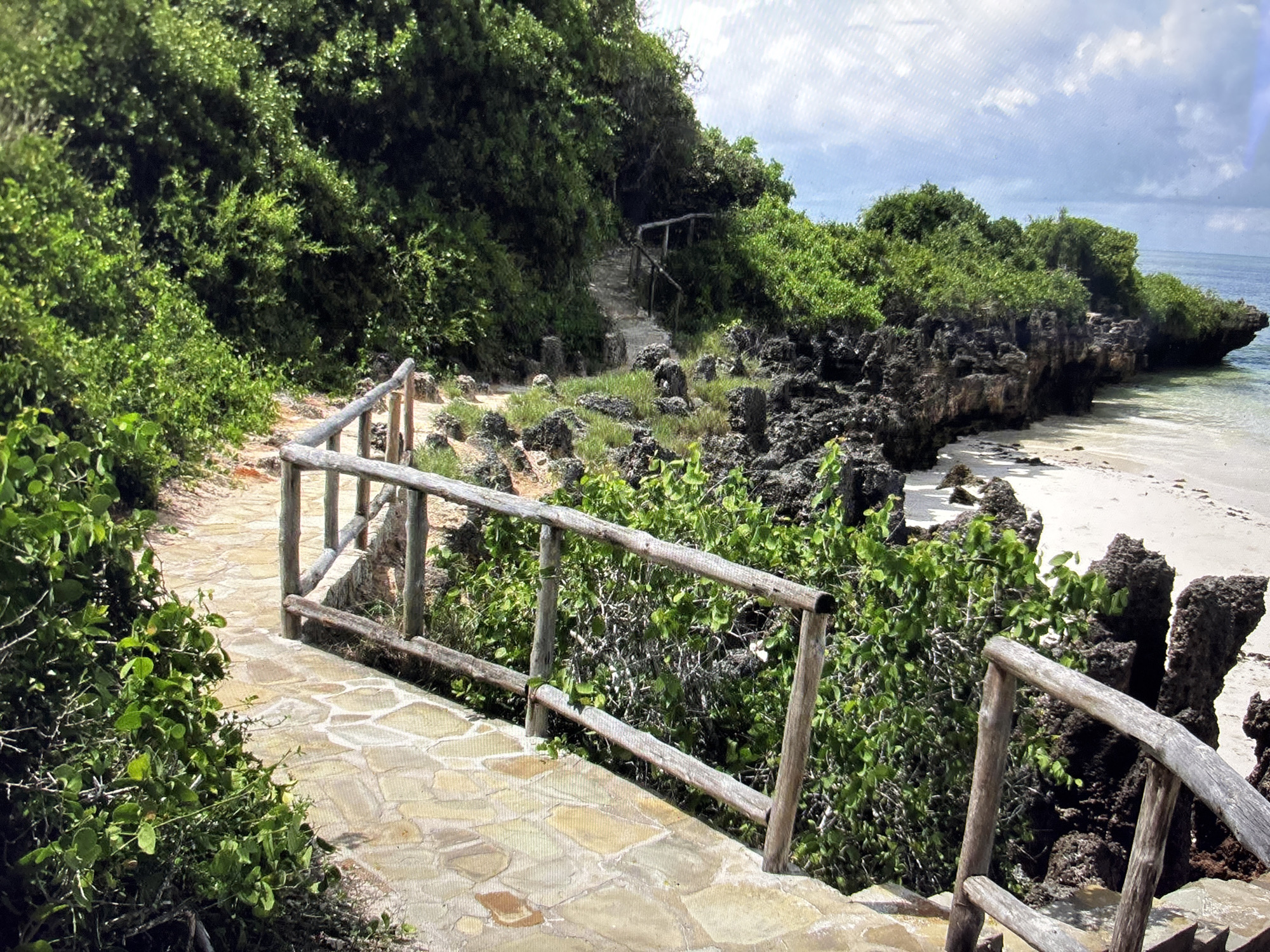 The image depicts a pathway with a wooden railing leading through lush green vegetation on a tropical island. The pathway is made of stone pavers and curves gently to the right. To the right of the pathway, there is a rocky shoreline with the ocean visible in the background. The sky is partly cloudy. The location is not in Sofia, Bulgaria; it appears to be a coastal area with natural scenery. There are no people or animals visible in the image.