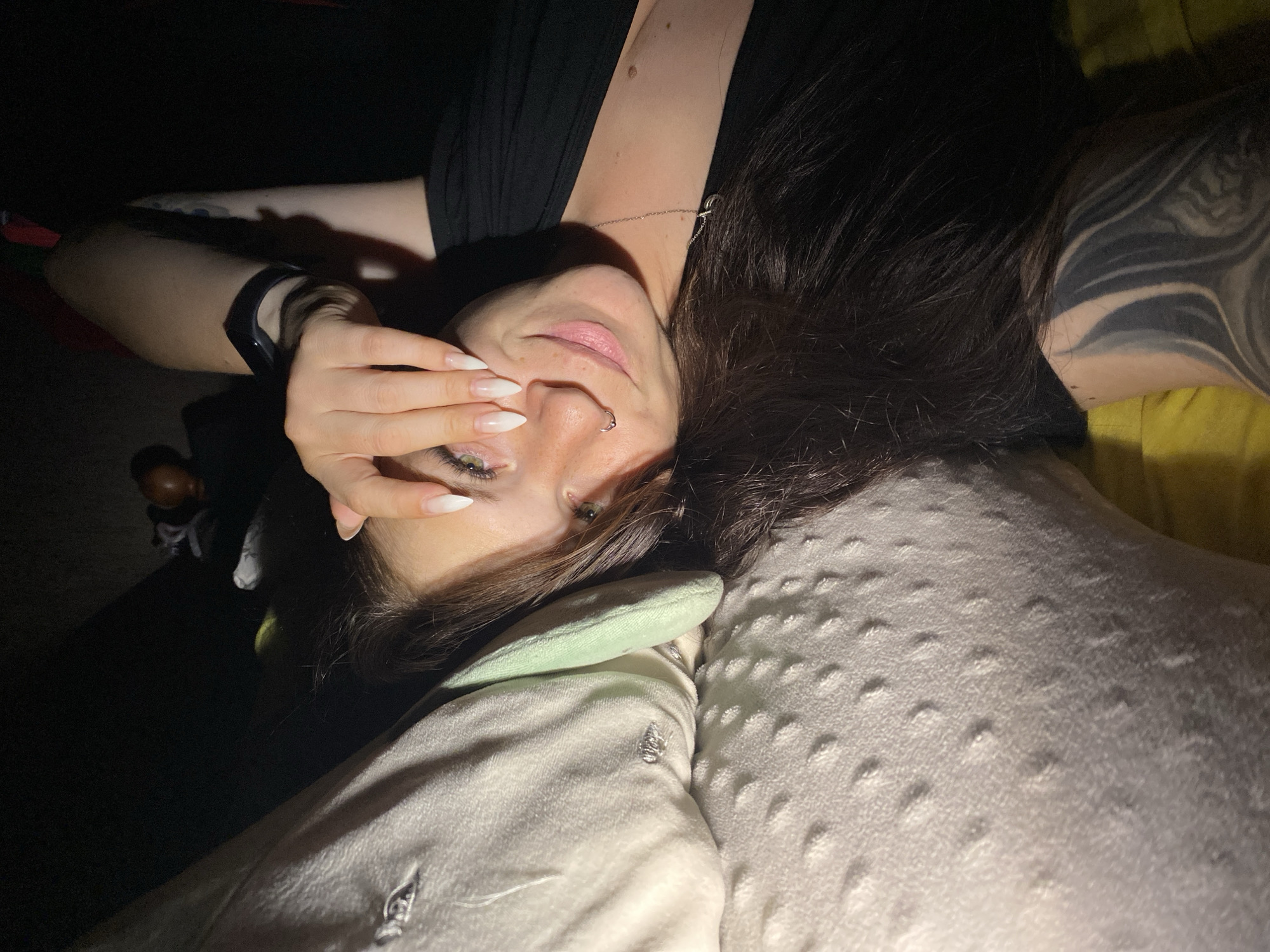 A woman with long dark hair and green eyes, wearing a black top and a silver necklace, lies on her side on a bed. Her right hand is covering her face, and she is wearing a fitness tracker on her left wrist. She has long, white, almond-shaped fingernails and a nose ring. A textured, light-colored pillow is to her left. The scene is dimly lit, with some areas of shadow. There is a tattoo visible on her right arm. The location is not specified in the image.