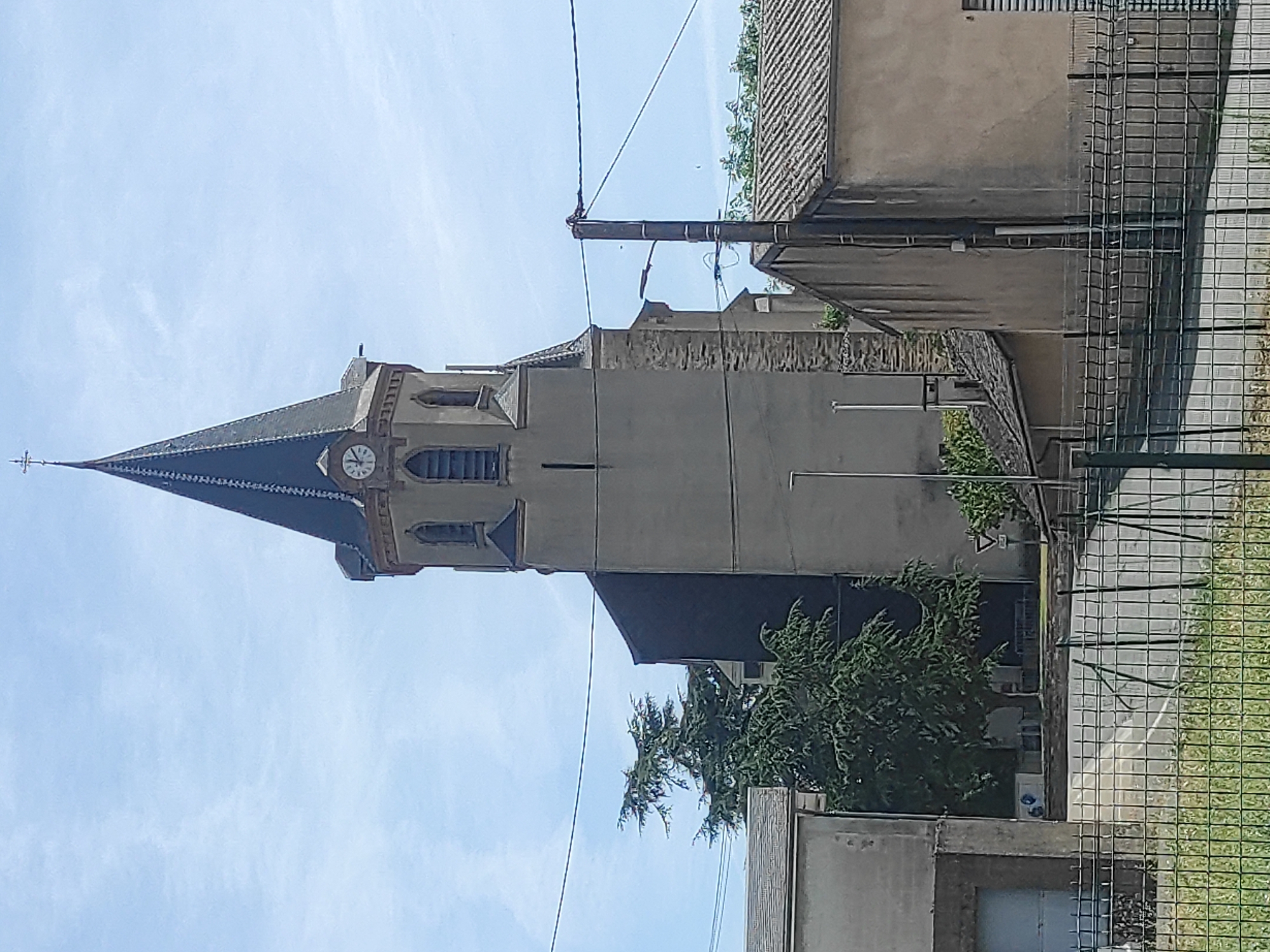 This is a vertically oriented, medium-shot photograph taken outdoors on a sunny day with a blue sky and scattered clouds. The main subject is a stone church tower with a dark, pointed roof and a white clock face. The tower has arched windows and decorative stonework. To the right of the church, a multi-story building with a tiled roof is visible, along with a chain-link fence and some greenery. The scene suggests a rural or small-town European setting, likely in Valence-d'Albigeois, France, given the context. There are no people visible in the image. The overall mood is peaceful and serene, with the bright sunlight casting shadows and highlighting the textures of the buildings.