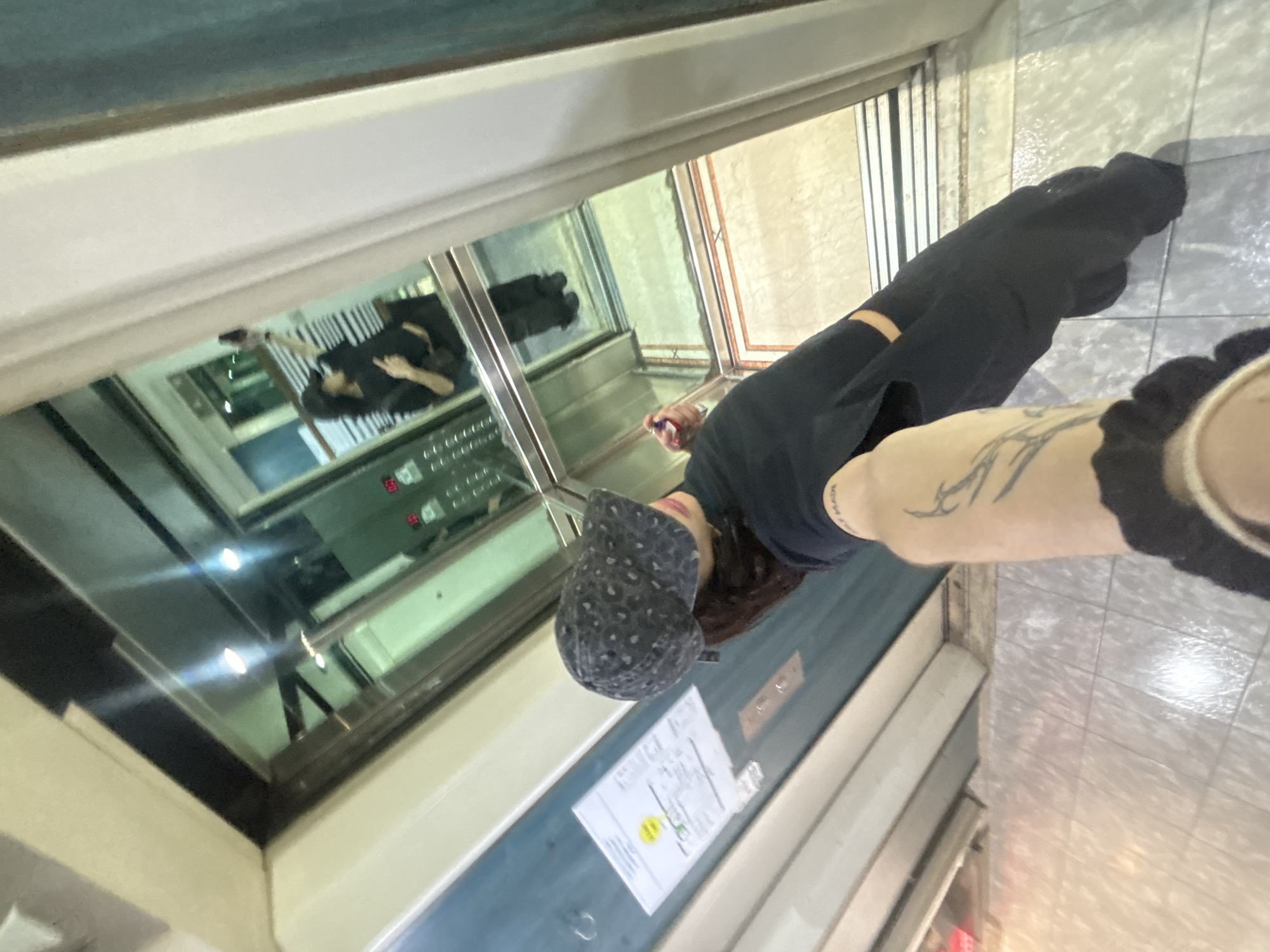 A distorted perspective captures a person in an elevator. The individual wears dark clothing and a patterned cap, with a tattoo visible on their arm. The elevator interior features a control panel and safety instructions. A reflection reveals another person inside the elevator. The surroundings include tiled flooring and wall paneling. The location is suggested to be in Buenos Aires, Argentina, based on unconfirmed details.