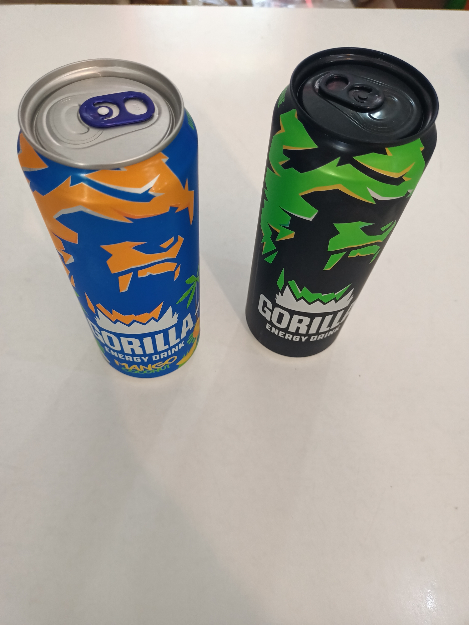 Two opened cans of Gorilla energy drink are positioned on a light-colored, reflective surface, likely a table. The can on the left is blue with orange and yellow abstract designs, and has a purple pull-tab. The can on the right is black with green and yellow abstract designs and has a black pull-tab. The lighting suggests an indoor environment. There are no people or any specific location markers that would suggest the city of Abay, Kazakhstan.