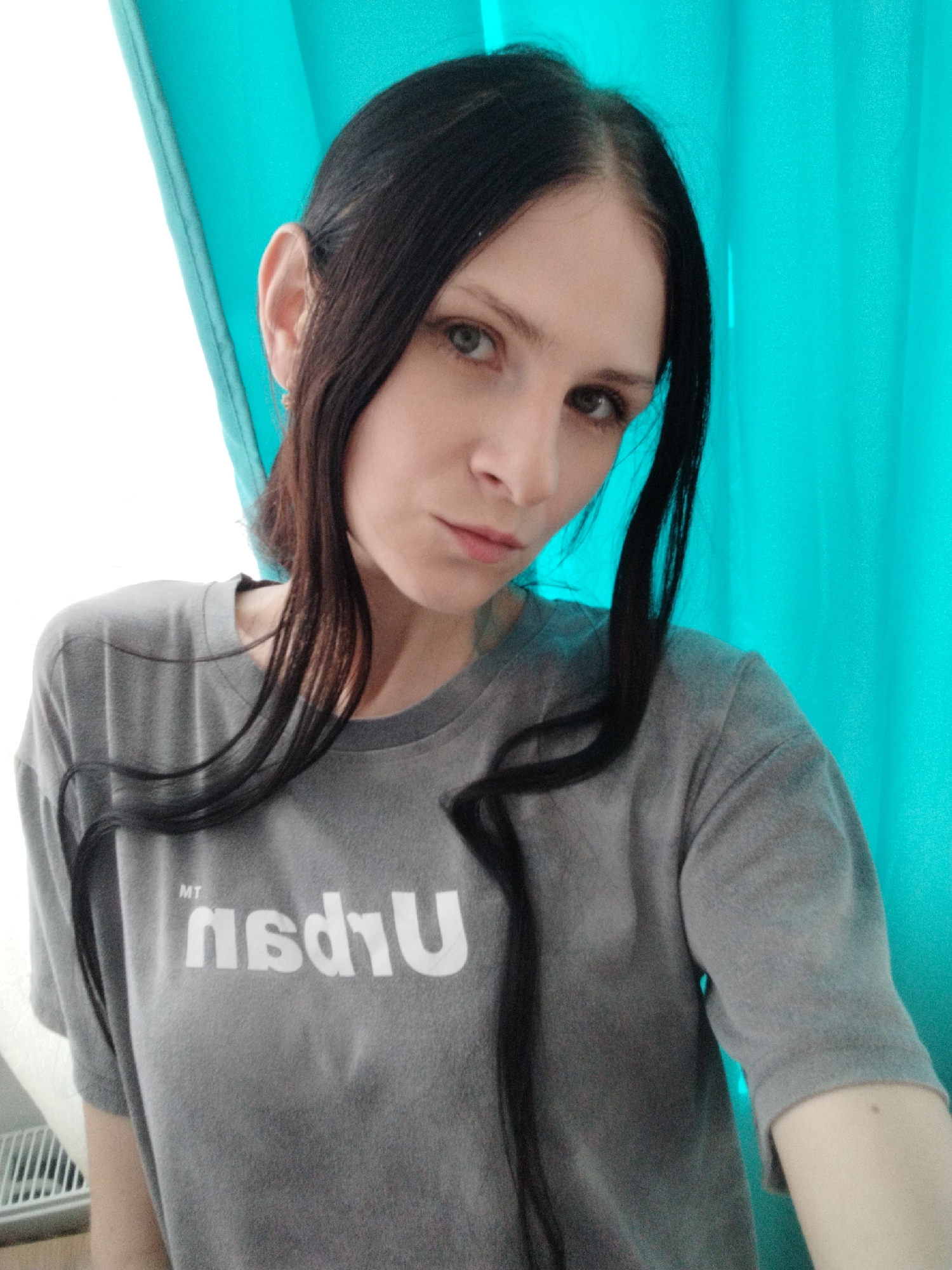 This is a close-up, waist-up, selfie-style photograph of a young woman with long, dark hair. She is wearing a plain grey t-shirt with white text on it that appears to be a logo. The text is partially obscured by her hair, but "MT" is visible above what looks like "nsdTU" or a similar reversed word. She has light skin and is looking towards the camera with a neutral expression. Her eyes are a light green or hazel color. Her hair is pulled back loosely, with strands falling around her face and shoulders.
The background consists of a bright teal-colored curtain or drape, which appears to be behind her. To the left of the image, there is a bright white area, possibly a window or another curtain, creating a strong contrast with the teal. The lighting suggests it is daytime, possibly indoors given the curtain. There are no other people or significant objects in the frame, focusing entirely on the subject and her attire. The overall mood is casual and direct.