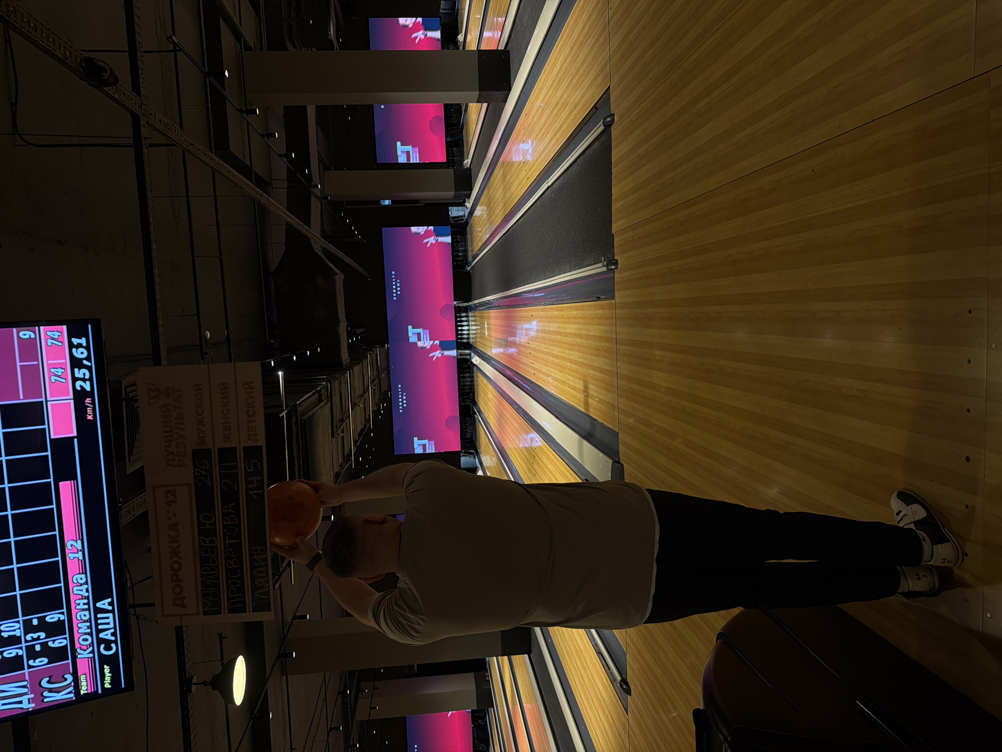 This image captures a person bowling inside a modern bowling alley in Izhevsk, Russia. The main subject, seen from behind, is a person wearing a light-colored t-shirt, dark pants, and bowling shoes, holding an orange bowling ball above their head, poised to release it onto one of the wooden lanes.
The scene is illuminated by artificial lighting, indicating an indoor, likely evening, setting. Polished wooden bowling lanes stretch into the distance, leading to sets of pins. Large digital screens above the pins display animated content and the "BROOKLYN BOWL" logo. The ceiling features exposed industrial-style structures and track lighting.
To the left, a digital scoreboard displays game information, including "KC", scores such as "9", "10", "6-3-", "6", "9", and identifies "Team Команда 12" (Team 12) with "Player САША" (Player SASHA). It also shows a score of "9" and "Km/h 25,61," indicating the ball's speed. A hanging wooden sign above the lanes lists "ДОРОЖКА 12" (Lane 12) and "ЛУЧШИЙ РЕЗУЛЬТАТ" (BEST RESULT), followed by records: "МУРАВЬЁВ Ю. 276 МУЖСКОЙ" (Muravyev Yu. 276 MALE), "ПРОСВРТОВА 211 ЖЕНСКИЙ" (Prosvertova 211 FEMALE), and "ЛЯПИН 145 ДЕТСКИЙ" (Lyapin 145 CHILD). The image conveys the atmosphere of a recreational bowling game.