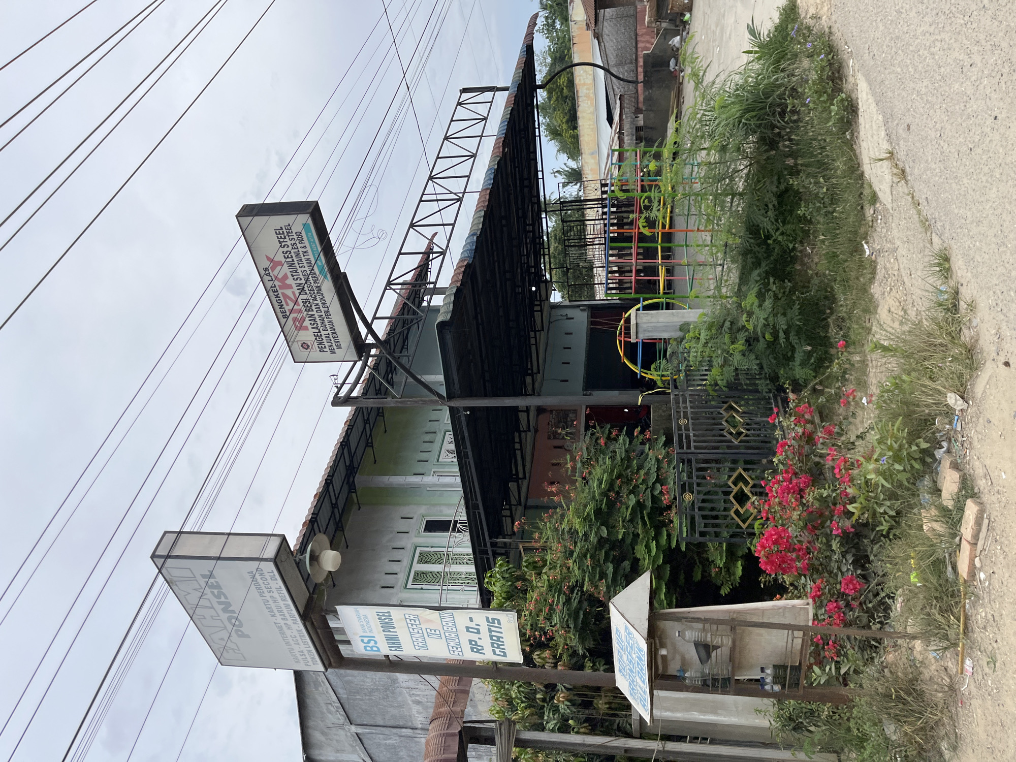The image shows a street view in Simpang Empat, Indonesia, featuring a multi-story building with several signs. A large white sign with black text at the top reads "FAHMI PONSEL" and lists services such as internet cards, pre-paid cards, and phone refills. Below this, a vertical banner with a white background and green/black text advertises "BSI FAHMI PONSEL TRANSFER KE SEMUA BANK RP. 0,- GRATIS" indicating free bank transfers. To the right, another large sign with a white background and red/black text advertises "BENGKEL LAS RIZKY" and lists services related to iron and stainless steel fabrication and accessories. A metal carport covers the front of the building, and colorful playground equipment is visible behind a fence. The environment appears to be a commercial area with some vegetation and overhead power lines. The ground is unpaved in some areas, and there is some litter present. No people are visible in the image.