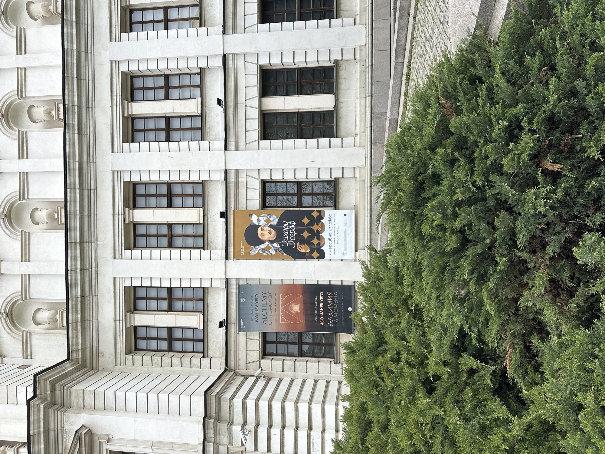 A grand, light-colored building, likely a cultural institution in Sofia, Bulgaria, is depicted from a slightly upward perspective during daytime. The architecture features classical elements, including prominent vertical pilasters, cornices, and decorative details like arched recesses and urn-like ornaments on the far left. The facade is punctuated by numerous rectangular windows with dark, multi-paned frames.
In the foreground on the right, a large, dense green conifer shrub or tree partially obscures the building, providing a natural contrast to the man-made structure. A paved area with light-colored stone slabs is visible behind the shrub.
Three vertical banners promoting art exhibitions hang on the building. The leftmost banner, featuring abstract gold patterns on a dark background, advertises "IVO ILIEV YETO ALCHEMY OF THE MOMENT," scheduled from "JUNE 2025-MAY 2026," curated by "MARTIN TASHKI" (МАРТИН КОСТАШКИ in Bulgarian). The adjacent banners, sharing a similar dark and gold aesthetic with a stylized portrait, promote a "ZAHARI ZOGRAPH IMMERSIVE EXHIBITION" (Захари Зограф Имерсивна изложба), running from "10.12.2025-30.01.2027." The text "НАЦИОНАЛНА" (National) is also visible, indicating a national venue.
The lighting is soft and even, suggesting either a clear day with indirect light or an overcast sky. The scene conveys a sense of cultural vibrancy in an imposing urban setting.