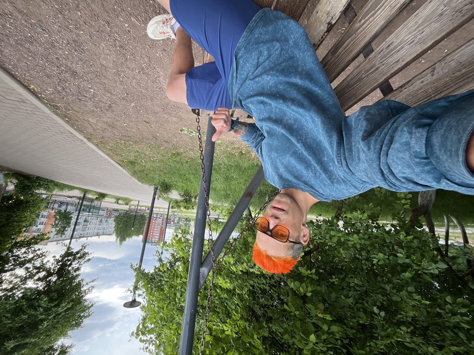 A man with orange hair and orange-tinted sunglasses, wearing a blue shirt and blue shorts, is sitting on a wooden bench in a park, facing the camera. He is next to a black swing set. A building can be seen in the background. The location is likely in the city of Chaikovsky, Russia.