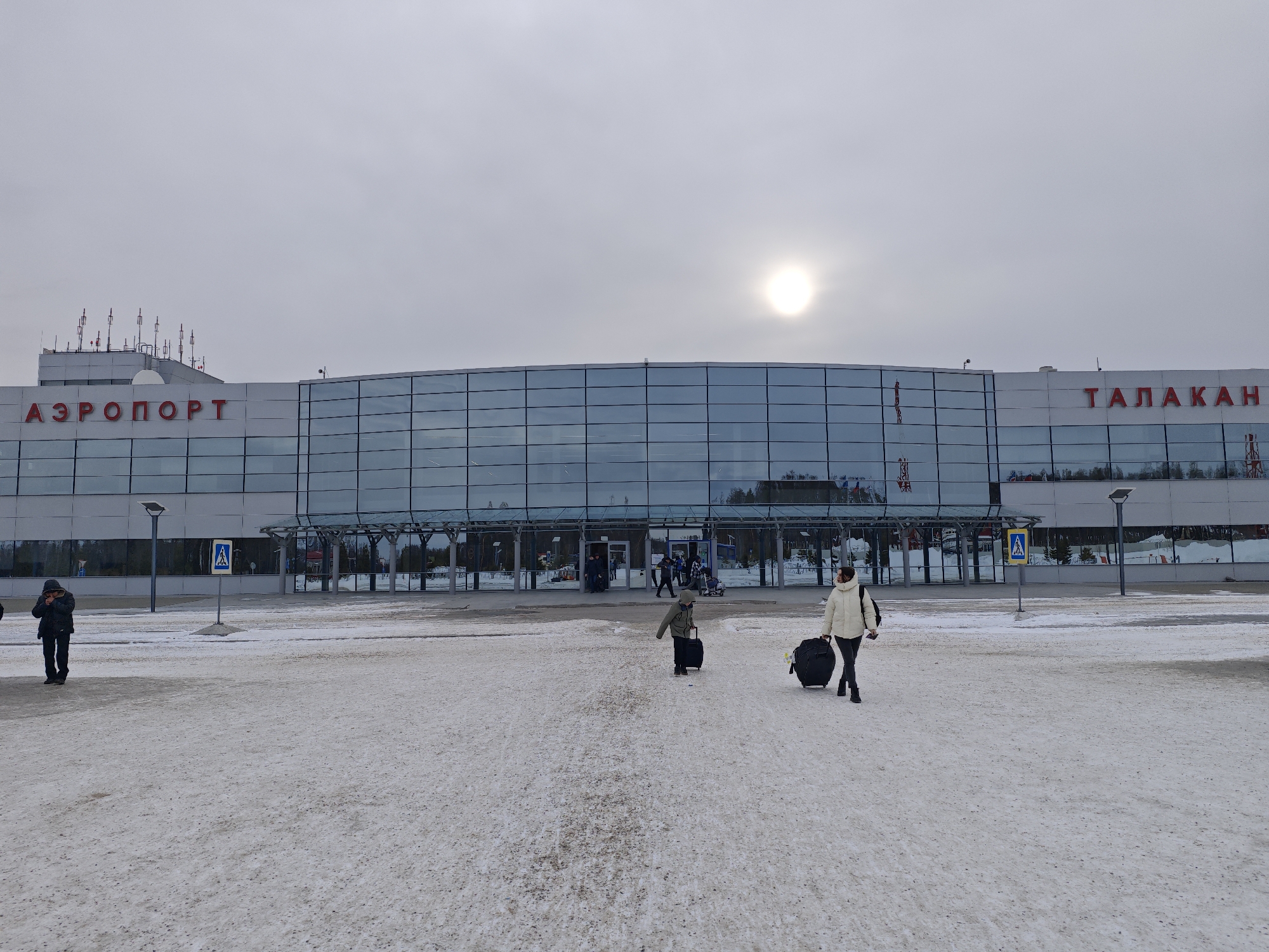 The image depicts the Talakan Airport in Russia. A large, modern building with the words "АЭРОПОРТ" and "ТАЛАКАН" on its facade in red lettering is prominent. The building has a primarily glass and steel structure. In the foreground, the ground is covered with snow. Several individuals are walking towards the airport entrance, pulling luggage. A woman in a white jacket is on the right, pulling a large suitcase. A child is walking in front of her, also pulling a suitcase. Another person is standing to the left, closer to the building. Streetlights and pedestrian crossing signs are visible. The sky is overcast.