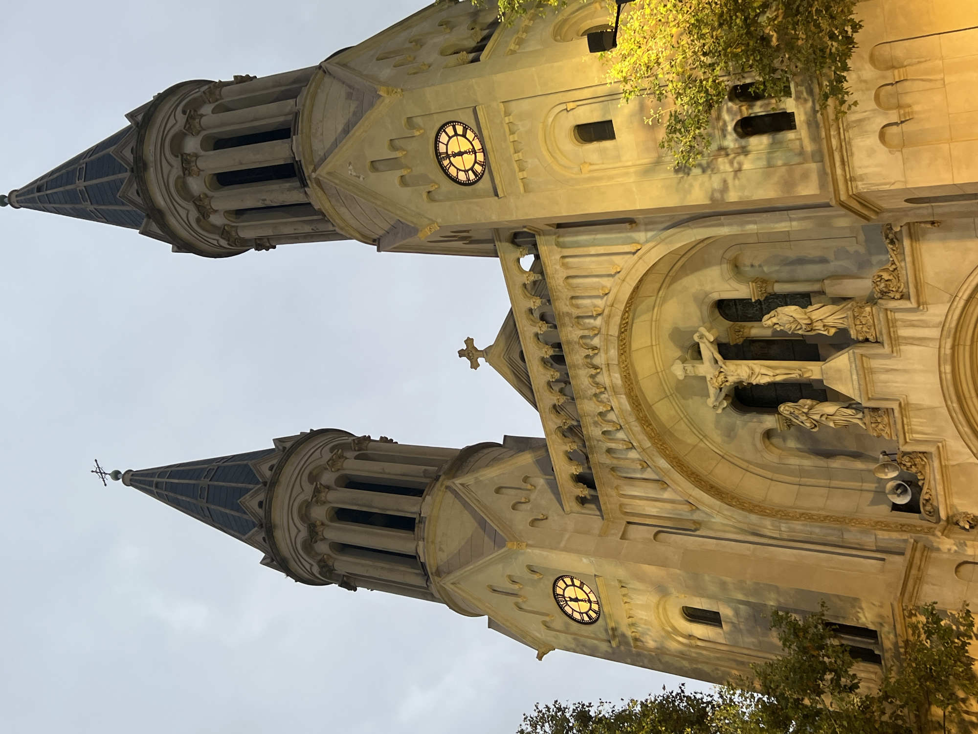 This is an exterior shot of the Basilica of the Holy Sacrament (Basílica del Santísimo Sacramento) in Buenos Aires, Argentina, captured at dusk or early evening, based on the sky. The angle is upward and slightly off-center, emphasizing the twin towers and facade. The towers are illuminated, highlighting the architectural details, including clocks, decorative stonework, and pointed spires. Some foliage is visible at the base of the building. The basilica is a prominent landmark in the city. The lighting gives the stone a warm, golden hue. No people are visible in the frame.