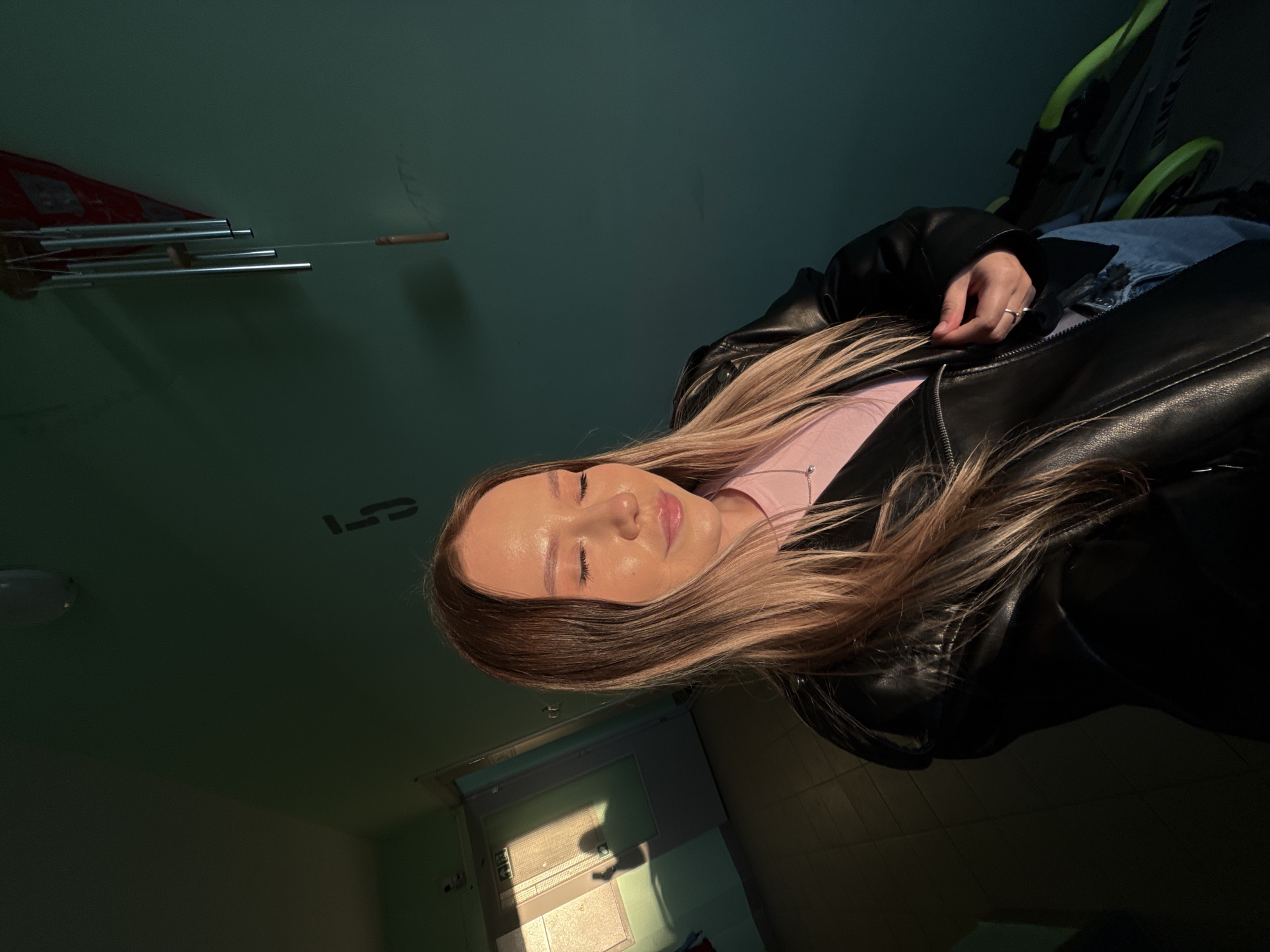 An indoor scene in the city of Chaikovsky, Russia, depicts a young woman with long, blonde and brown ombre hair, standing in what appears to be a hallway or entryway. Her eyes are closed, and her lips are slightly parted in a faint smile. She wears a light pink top beneath an open black leather jacket, with blue jeans partially visible in the lower right. Her left hand is positioned near the zipper of her jacket, displaying a ring on her left ring finger. A strong, warm light source from the left illuminates the left side of her face and hair, creating highlights and casting shadows. The wall behind her is a dark teal or emerald green, featuring a stenciled black numeral "5" in the upper middle. To the upper right, a wind chime with silver tubes and a wooden clapper hangs from the ceiling. In the background to the left, a doorway leads to another area where sunlight casts a grid pattern from a window with blinds, as well as the distinct profile shadow of a person, onto a surface. The floor is covered with dark tiles. A portion of a bicycle with a bright green frame is visible in the lower right, partially obscured by the woman.