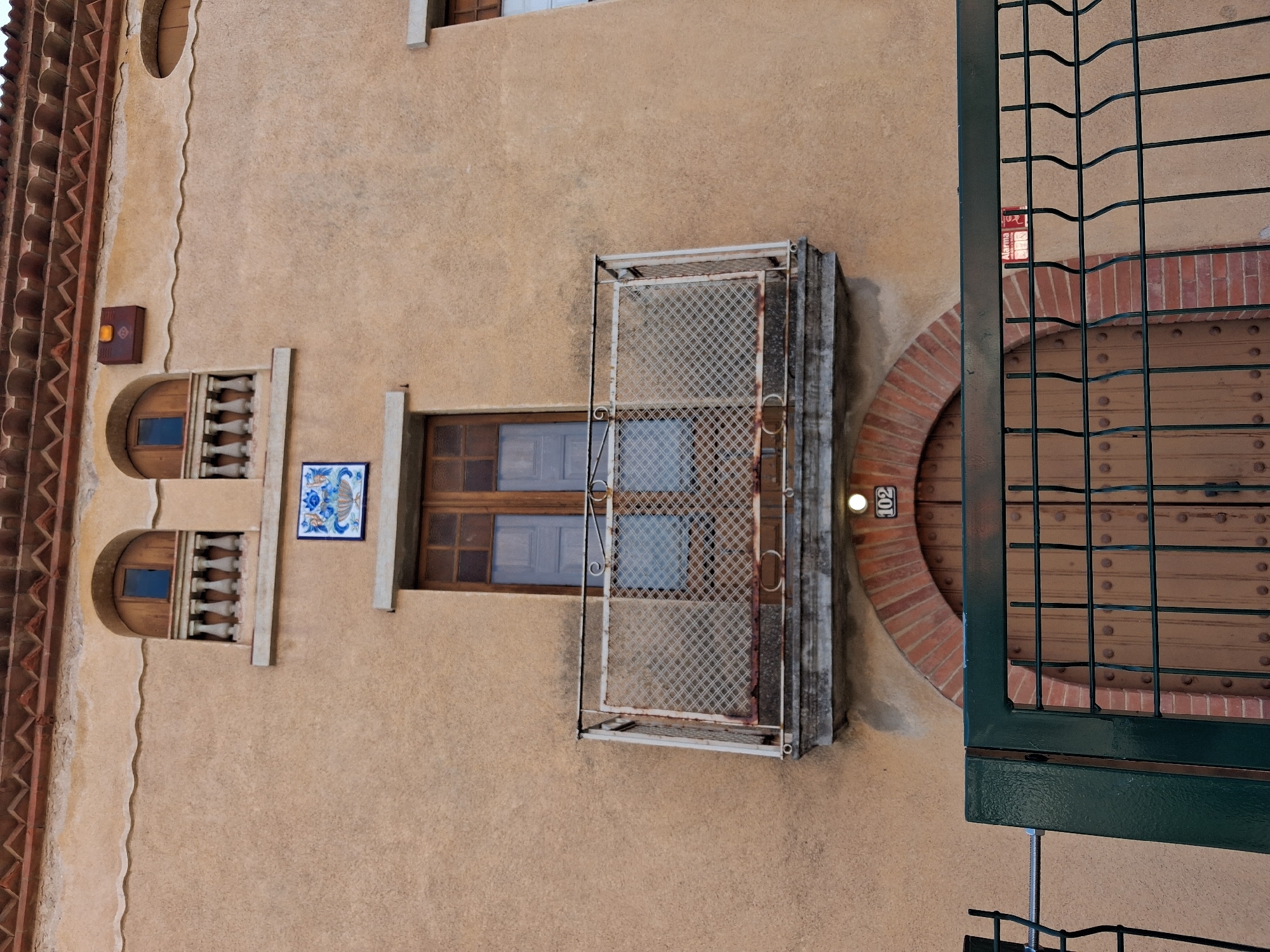 The image shows the facade of a building in Vilablareix, Spain. The building has a light brown stucco exterior and a red tile roof with decorative eaves. On the second story, there are two arched windows with balconies, each with a balustrade and a metal railing. A decorative blue and white tile is mounted between the windows. Below the windows, a small balcony extends from the building, supported by corbels and featuring a decorative metal railing with a diamond pattern. A wooden door with metal studs is visible below the balcony, framed by a red brick arch. The number "102" is displayed above the door. A dark green metal fence with a wavy top is in the foreground, partially obscuring the lower part of the building. An "Alarma" (alarm) sign is visible on the upper right side of the building.