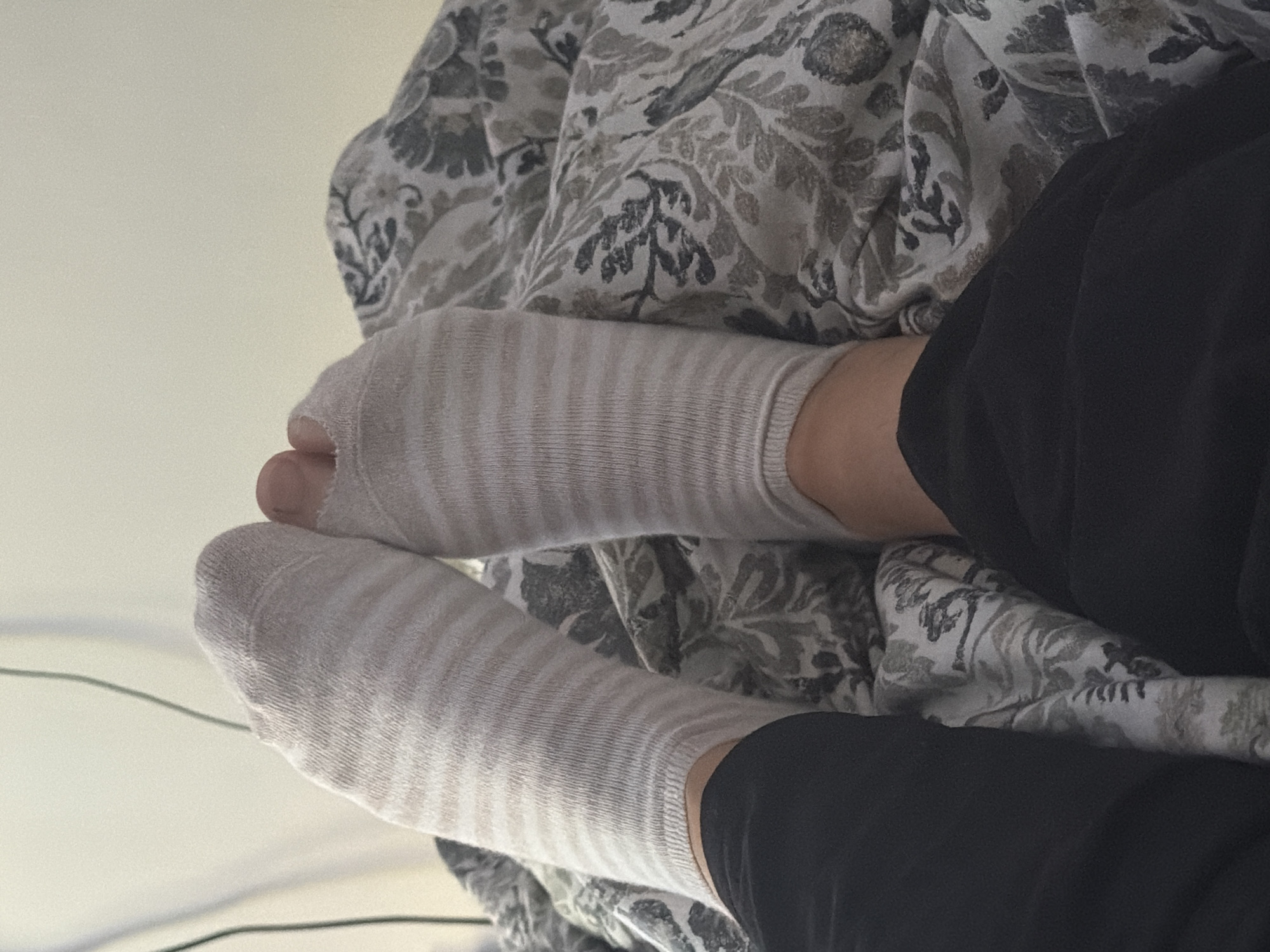 A close-up, elevated perspective depicts two human feet, clad in light-colored, horizontally striped ankle socks, resting on a patterned textile surface, likely a blanket or duvet. The sock on the right foot has a visible tear or hole at the toe, exposing the large toe. The individual's lower legs are partially visible, covered by black fabric, consistent with pants or leggings.
The patterned textile features an intricate design of leaves and flora in shades of gray, white, and muted earthy tones. In the upper left background, a plain, light-colored wall or surface is visible, against which thin, dark electrical wires or cables are hanging. The scene is indoors, indicating a relaxed setting.
The image contains no discernible features, landmarks, or contextual information to indicate its geographical location in Maidstone, United Kingdom, or any other specific city or country.