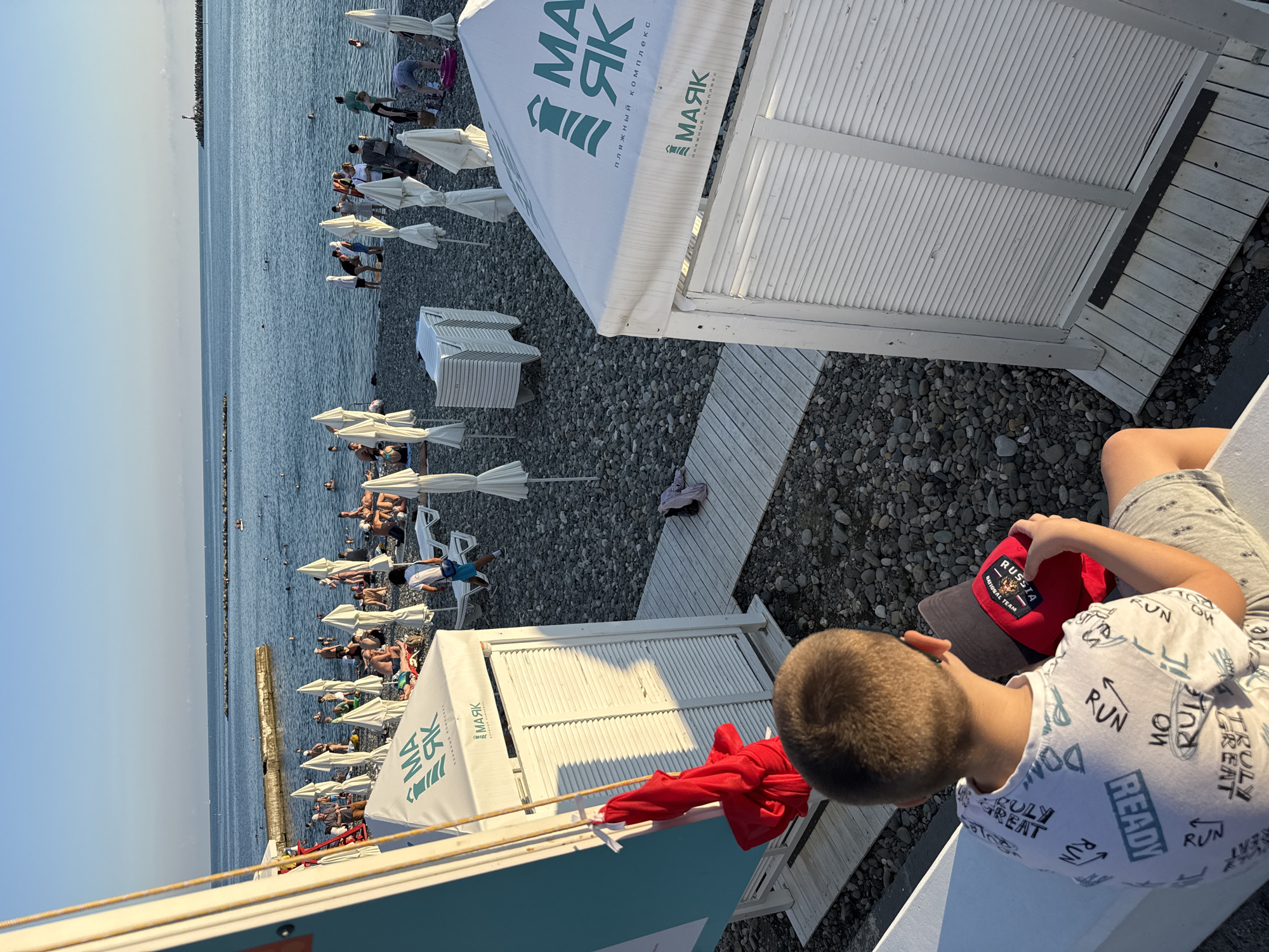 The image shows a sunny day at a crowded pebble beach in Sochi, Russia. The sea is visible in the background with several people swimming and sunbathing. There are white umbrellas and beach tents set up on the shore. In the foreground, a young boy with short brown hair is sitting on a white ledge, facing away from the camera. He is wearing a white t-shirt with various words and graphics printed on it, and grey shorts. He is holding a red hat with a patch on it. The beach has a wooden walkway and is covered in grey pebbles. The overall scene depicts a typical summer day at a popular seaside resort.