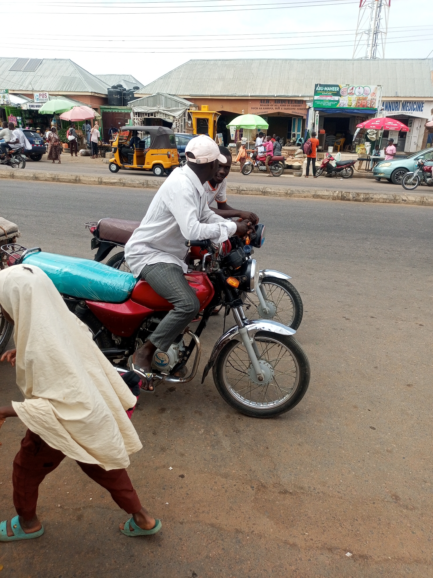 The image depicts a street scene in Tempure, Nigeria. In the foreground, two men sit on separate motorcycles parked on the side of the road. The man closest to the camera is wearing a light-colored cap, a white long-sleeved shirt, and grey pants, and he is wearing sandals. A second man is sitting on another motorcycle behind him. A person wearing a tan covering is walking in front of the motorcycles. In the background, there are shops with awnings and signs, including one that reads "ABU-MAHEER GENERAL VETERINARY". There are people walking and motorcycles parked along the street in front of the stores. A yellow "keke" or auto-rickshaw is visible. A tower is visible on the right behind the buildings.