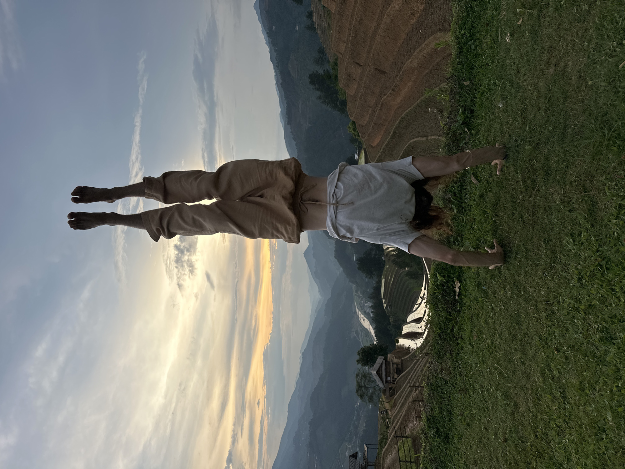 An individual performs a handstand on a grassy area, set against a breathtaking mountainous landscape. The person is viewed from behind, wearing loose, light-colored pants and a light t-shirt. Their arms are extended downwards, hands on the grass, while their legs are held straight up towards the sky, showcasing balance and strength.
The setting is a vast valley characterized by multiple layers of terraced rice fields, some of which are filled with water, reflecting the warm light. These terraces cascade down steep hillsides, creating intricate patterns across the landscape. Beyond the cultivated fields, hazy mountains stretch into the distance under a expansive sky.
The time of day appears to be either sunset or sunrise, as indicated by the soft golden light radiating from behind clouds in the sky. Patches of clear blue sky are also visible alongside more textured cloud formations. The overall scene suggests a tranquil yet powerful moment, combining human athleticism with the natural beauty of the Mù Cang Chải region in Vietnam. No visible text is present in the image.