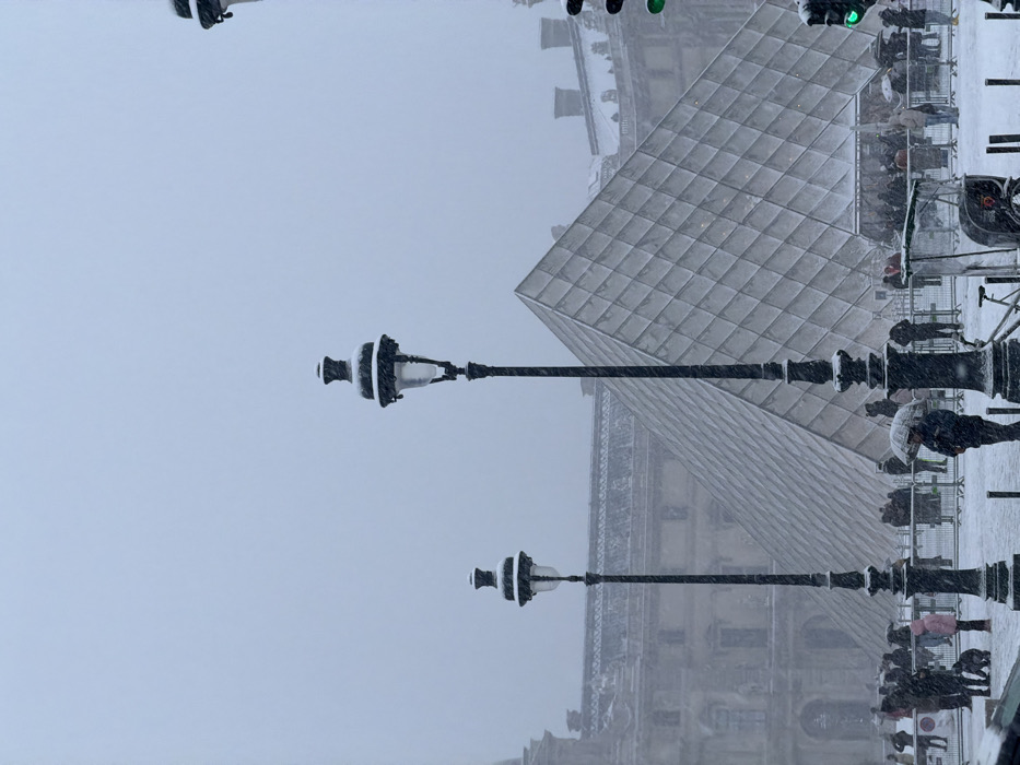 The image depicts the Louvre Pyramid in Paris during a snowstorm. Two ornate lampposts, partially covered in snow, stand in the foreground. Behind them, the pyramid's glass panels are visible through the falling snow. People with umbrellas can be seen in the background, suggesting they are waiting in line or passing by the iconic structure. The overall atmosphere is cold and overcast.