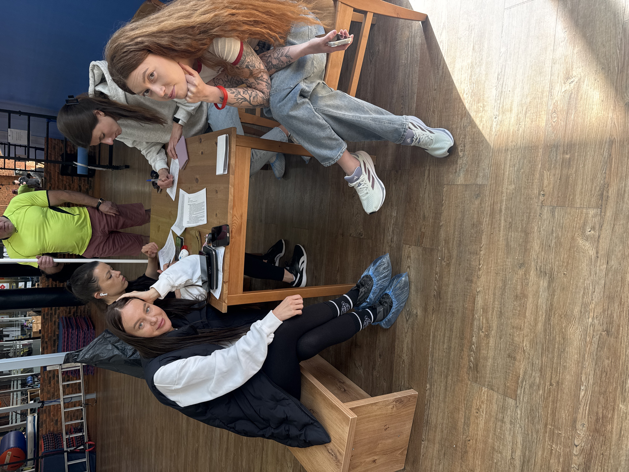 The image depicts a group of four women and one man, indoors, likely in a gym or sports-related facility. The location is Chaikovsky, Russia. Two women are seated at a wooden table, apparently filling out forms or documents. One of them, with long red-brown hair and tattoos on her arm, is holding a phone and looking at the camera. Another woman is sitting on a small wooden shelf or bench, also looking at the camera. The woman at the table has a pen in her hand, as if she is filling in a document. In the background, the gym is visible, with equipment like weights, ropes, and punching bags. One man wearing a neon green shirt is standing in the background. The floor is made of wooden planks. All the women are wearing casual clothes like jeans and hoodies, with the one on the bench wearing leggings and a puffer vest.