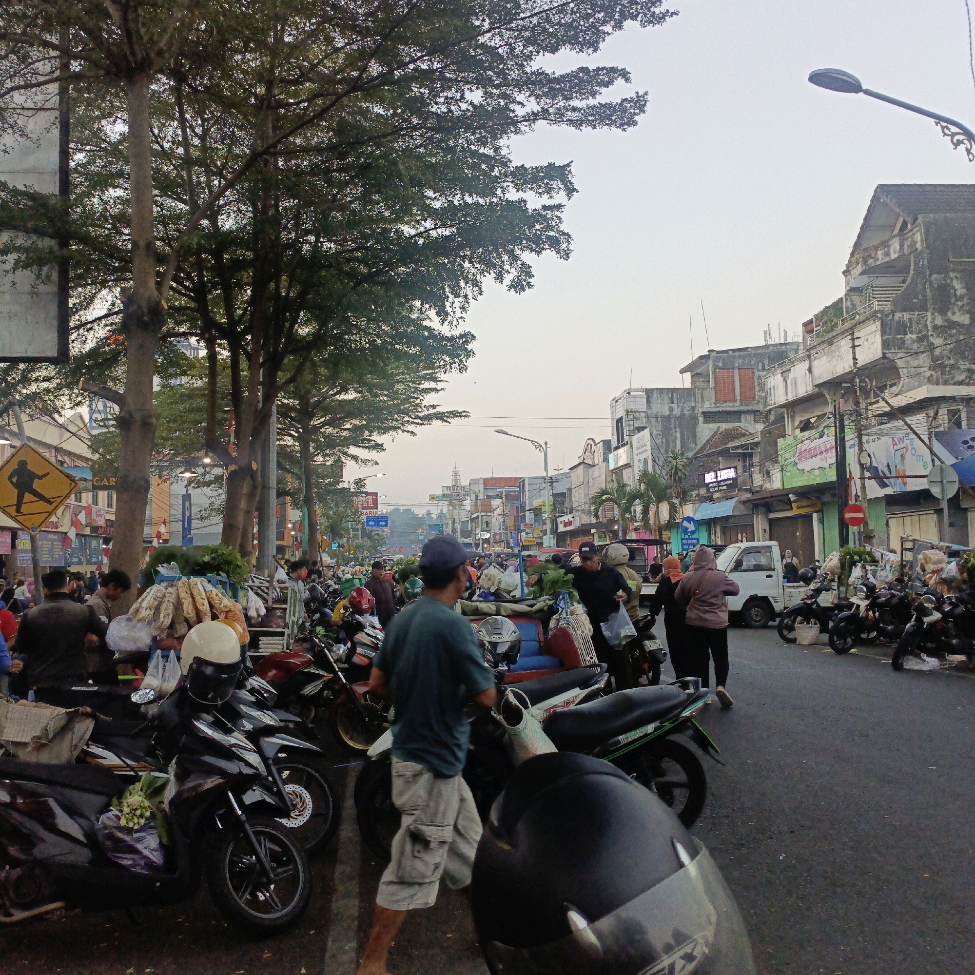 The image depicts a busy street scene in Salatiga, Indonesia, likely a market area. The foreground is dominated by several parked motorcycles, some with helmets and produce bags on them, indicating a lively commercial environment. In the mid-ground, a number of people are walking, shopping, and interacting. Some are carrying items, and others appear to be vendors. The background shows buildings with a somewhat worn architectural style, typical of older urban areas, and trees lining the street, suggesting a mature urban landscape. The sky is overcast, hinting at either early morning or late afternoon. A yellow diamond-shaped pedestrian crossing sign is visible on the left, adding to the urban setting. The overall impression is one of a bustling everyday scene in a city that blends commercial activity with a somewhat traditional urban aesthetic.