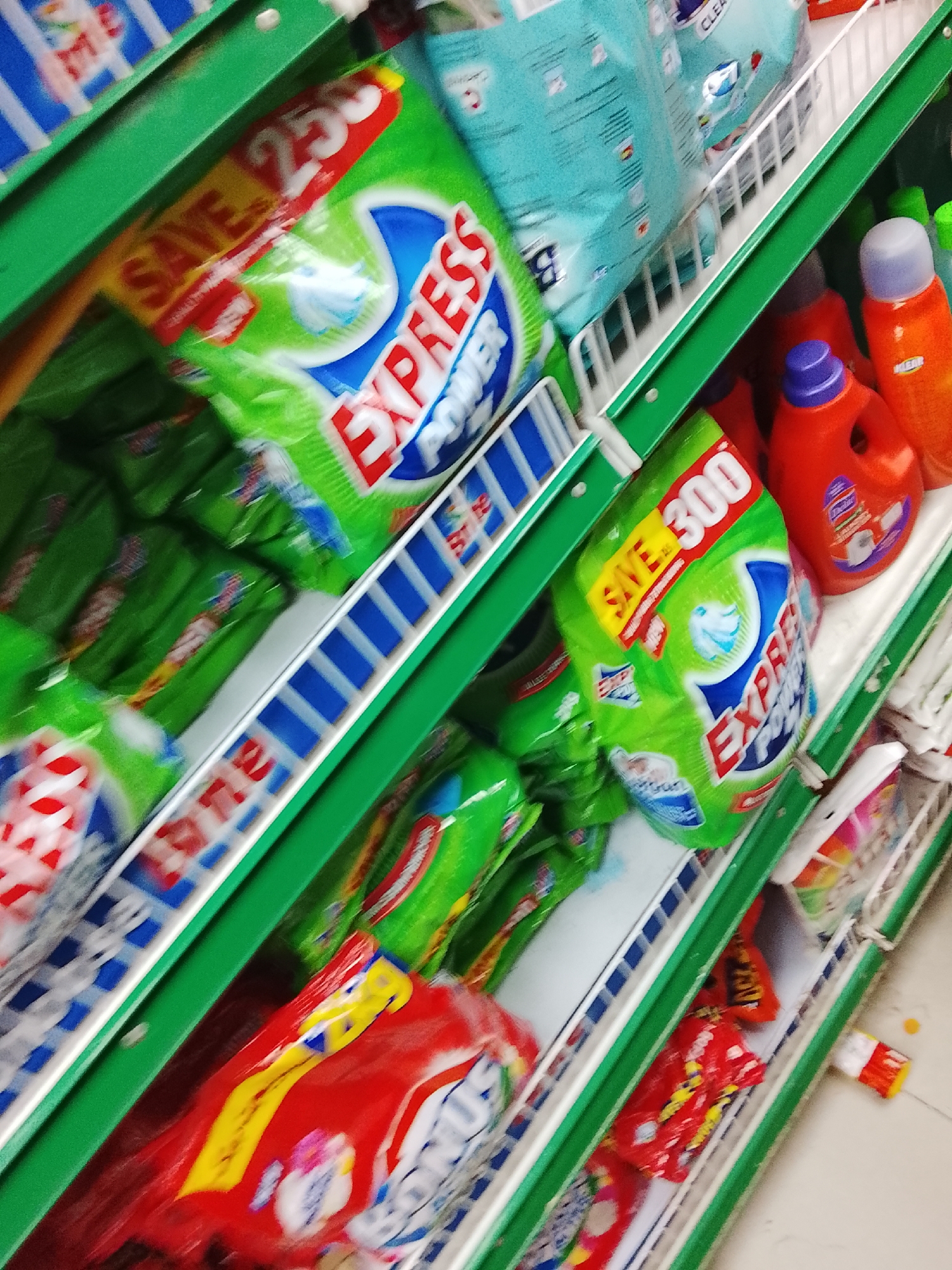 A slightly blurry, angled close-up shot of a grocery store shelf in Gulgasht Colony, Pakistan. The shelves are green and white with blue accents. They are stocked with various brands of detergent and cleaning products, including packages of Express Power detergent in green and blue packaging, as well as red and yellow detergent packages, and bottles of orange cleaning liquid. Discount signage is visible on some of the items. The floor is light-colored.