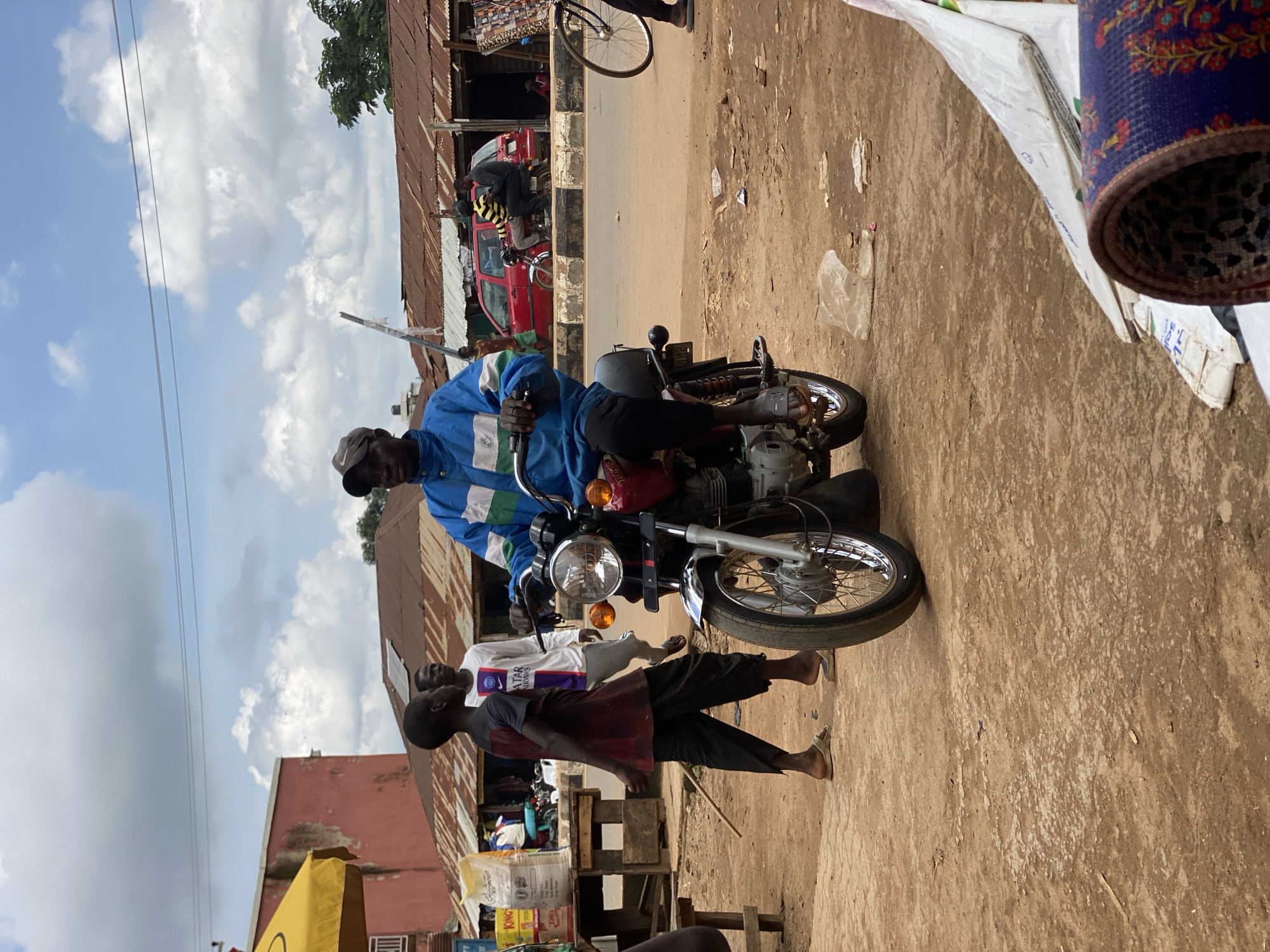 A man in a blue and white jacket and a dark cap rides a red motorcycle with silver trim. He is on a dirt road in front of several buildings with corrugated metal roofs. To his left, two boys are walking; one is wearing a dark red shirt and black pants, and the other is wearing a white shirt with purple lettering. In the background, a red car is parked. The sky is cloudy. The scene appears to be a market or busy street in Kafanchan, Nigeria.