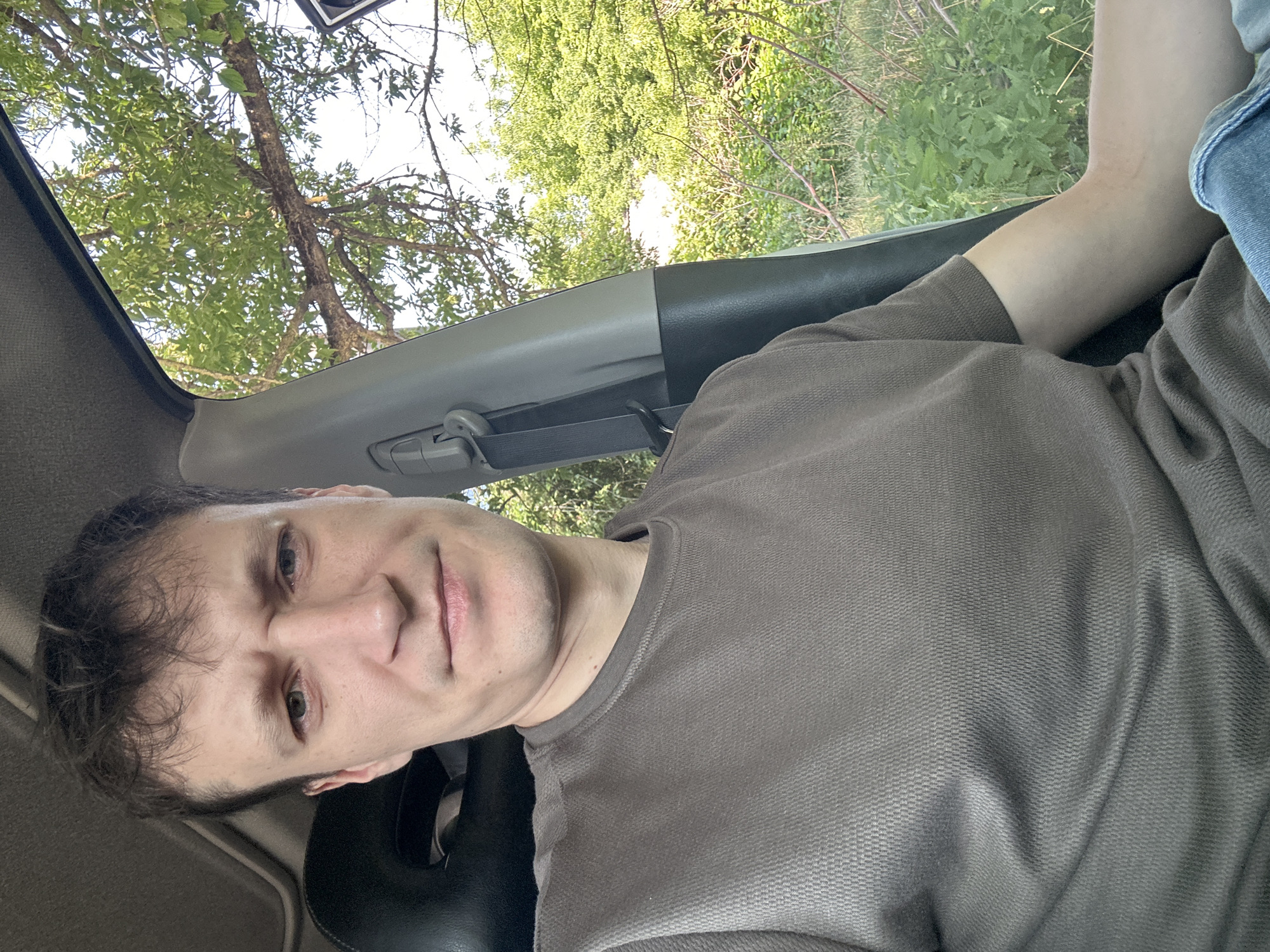 A medium shot captures a man sitting in the driver's seat of a vehicle, looking directly at the camera. He has light skin, short brown hair, and is wearing a brown t-shirt. His expression is neutral, with his eyebrows slightly furrowed. The interior of the car is visible, including the headrest of the passenger seat and part of the dashboard. Through the car window, lush green trees and foliage are visible, suggesting an outdoor, natural setting. The lighting indicates it is daytime, likely with sunlight filtering through the leaves. The specific location of Makiv, Ukraine, is not discernible from the visual information provided in the image.