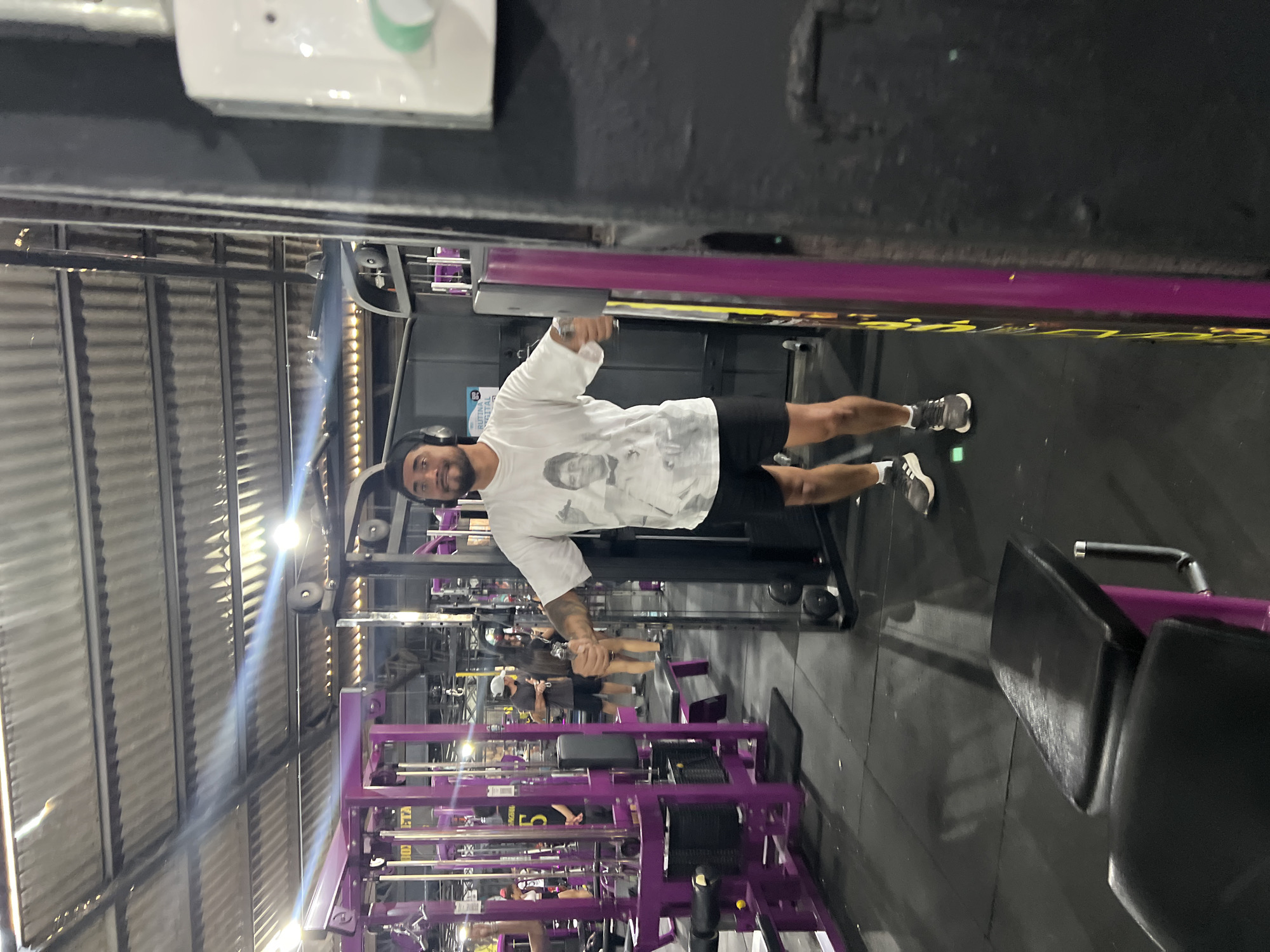 Arica, Chile: Interior view of a Planet Fitness gym, showing a man wearing a white t-shirt, black shorts and headphones working out on gym equipment. The gym has purple equipment and dark flooring. Other people are visible in the background, using various machines.
