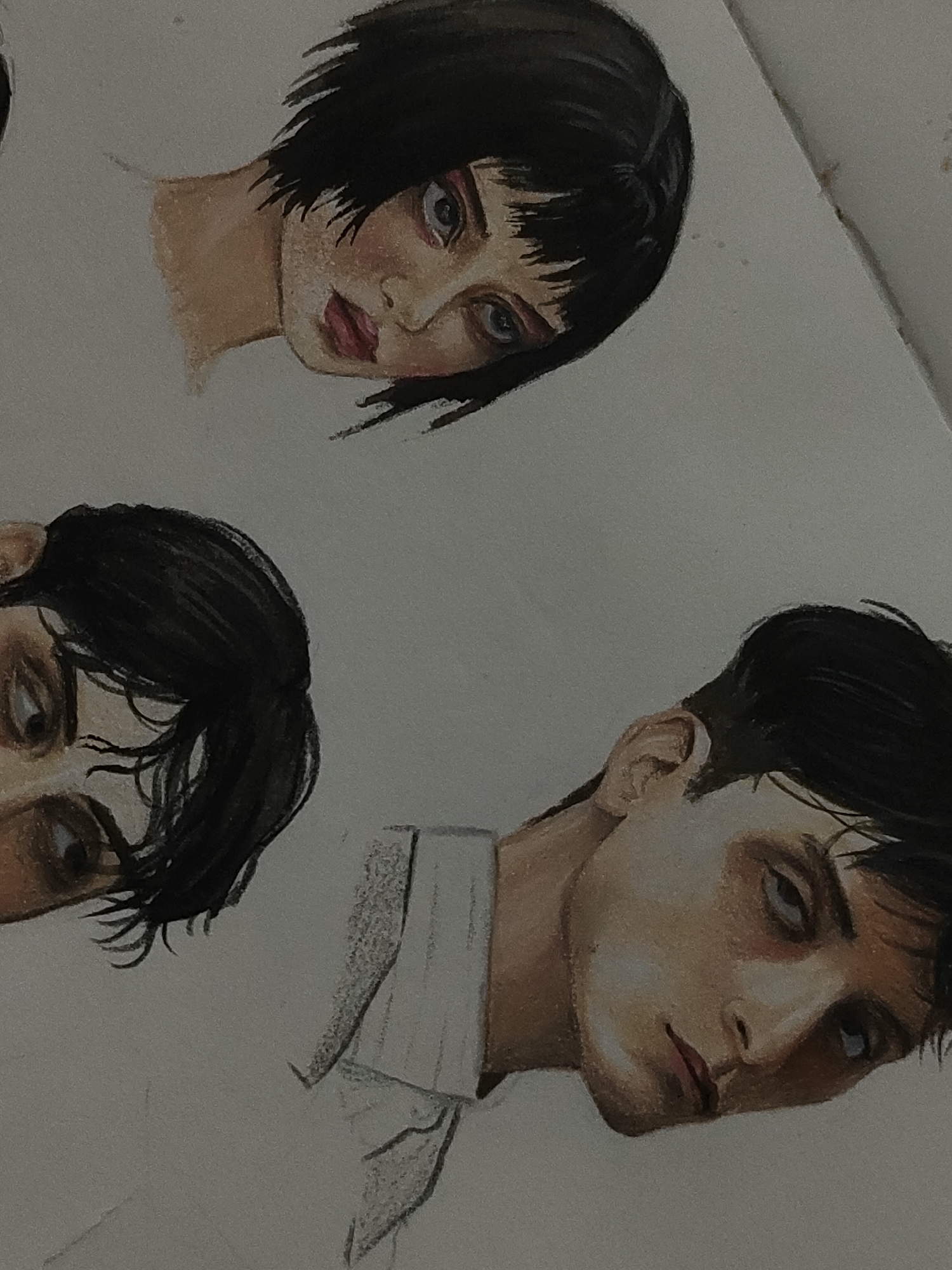 The image displays a drawing on a light-colored paper surface, featuring three distinct human faces rendered in a stylized, semi-realistic manner. The paper is oriented diagonally, with the subjects appearing to be tilted.
The uppermost subject is a female with short, choppy black hair and bangs. Her face is pale with a subtle flush on the cheeks and reddish, slightly parted lips. Her eyes, depicted with dark liner, show blue irises and gaze slightly upward. Her neck is visible, tapering down to an uncolored area.
Below and to the left of the top subject, a partial female face is visible, showing one dark eye looking downward, framed by dark hair and shading.
The lowest subject is a male face positioned to the right of the partial face. He has short, dark hair swept back from his forehead. His complexion is pale with prominent cheekbones and dark shading around his downward-gazing eyes, which have dark irises. His lips are thin and tinged with red. A portion of his right ear is visible, and he wears a white collared shirt, indicated by the collar on his left shoulder.
The drawing technique appears to involve colored pencils or a similar medium, showcasing visible texture in the shading and coloring of skin, hair, and facial features. The background is a uniform plain white, indicating an artistic rendering on paper rather than a photograph of a physical environment. There are no contextual elements within the image that allow for the determination of a specific geographical location, such as Davao City, Philippines.