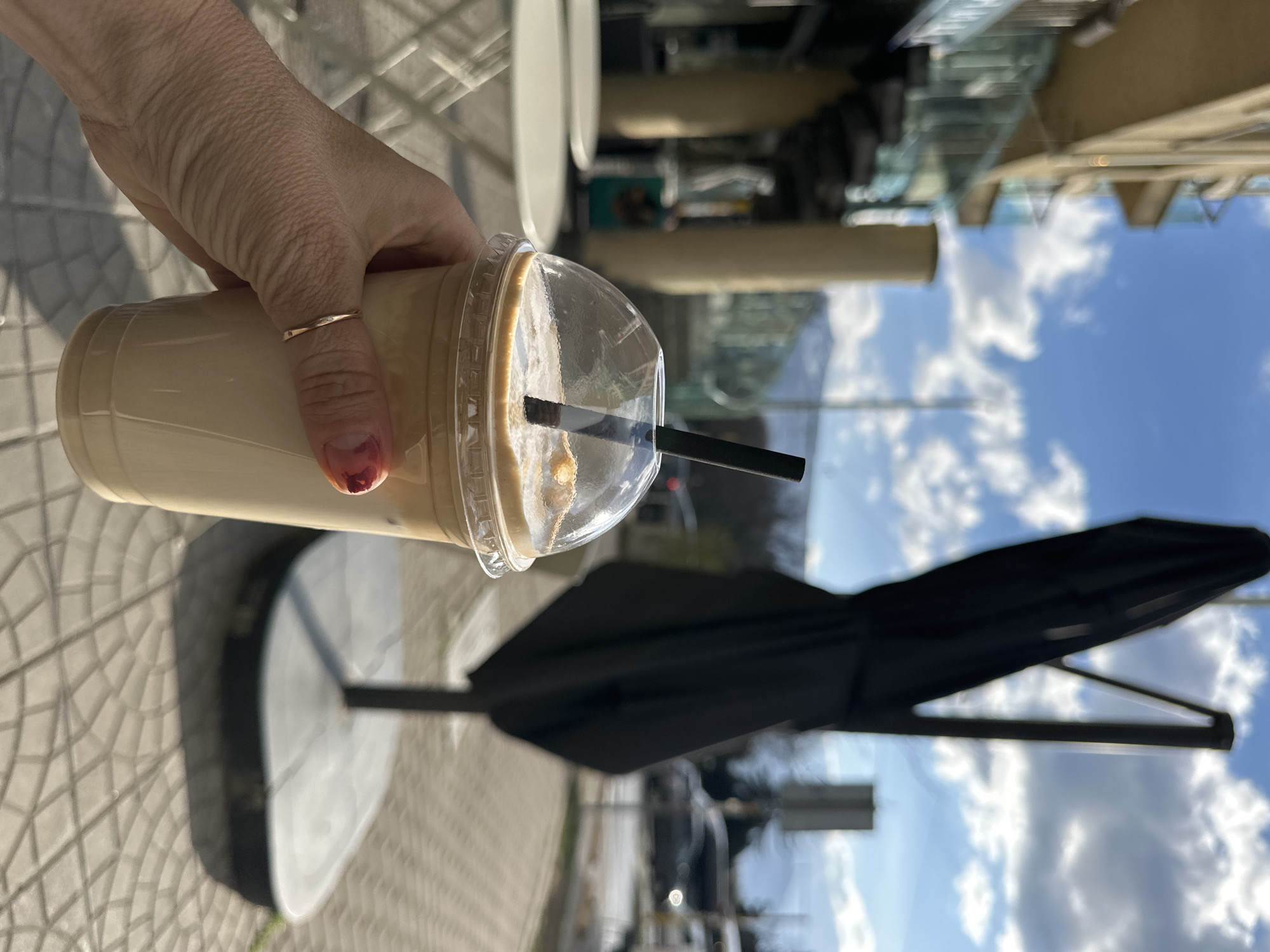 A human hand, with a thin gold-colored ring on the ring finger and chipped red nail polish on the index finger, holds a clear plastic cup containing an iced, light-brown beverage with a black straw and dome lid. The scene is an outdoor cafe situated on a paved sidewalk in Sofia, Bulgaria. The ground is composed of light-colored, interlocking paver stones. In the blurred background to the left, a multi-story building with light-colored facades and balconies featuring clear glass railings is visible. To the right, a large, black, folded outdoor umbrella stands on a dark base. Beyond the immediate cafe area, a blurred street with several vehicles is visible. A prominent mountain range is discernible in the distant background beneath a blue sky with scattered white clouds. Out-of-focus white outdoor cafe tables and chairs are visible to the left of the hand holding the beverage. The environment is brightly lit by natural sunlight, indicating daytime.