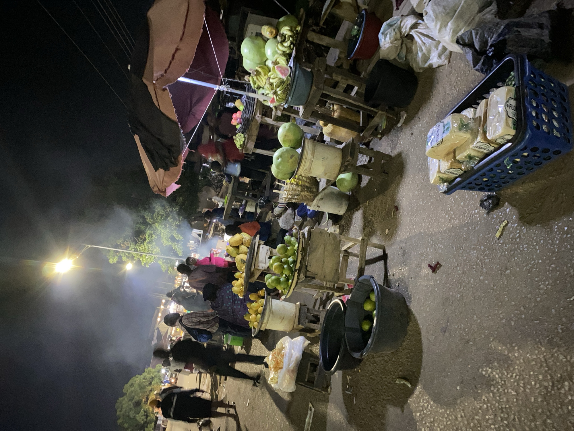 The image depicts a bustling street market scene at night, likely in Bukuru, Nigeria. Several vendors are displaying fruits and food items on makeshift wooden stands. The stands are topped with baskets and buckets containing fruits like bananas, watermelons, and mangoes.
People are gathered around the stalls, interacting with the vendors, presumably buying and selling goods. The light source seems to be a street lamp, illuminating the immediate area and casting shadows. Smoke or haze is visible, potentially from cooking or grilling.
A blue plastic basket filled with packaged food items sits near the edge of the street. The ground appears to be paved, and there's some litter visible. Overhead, a canopy provides some shelter for vendors.