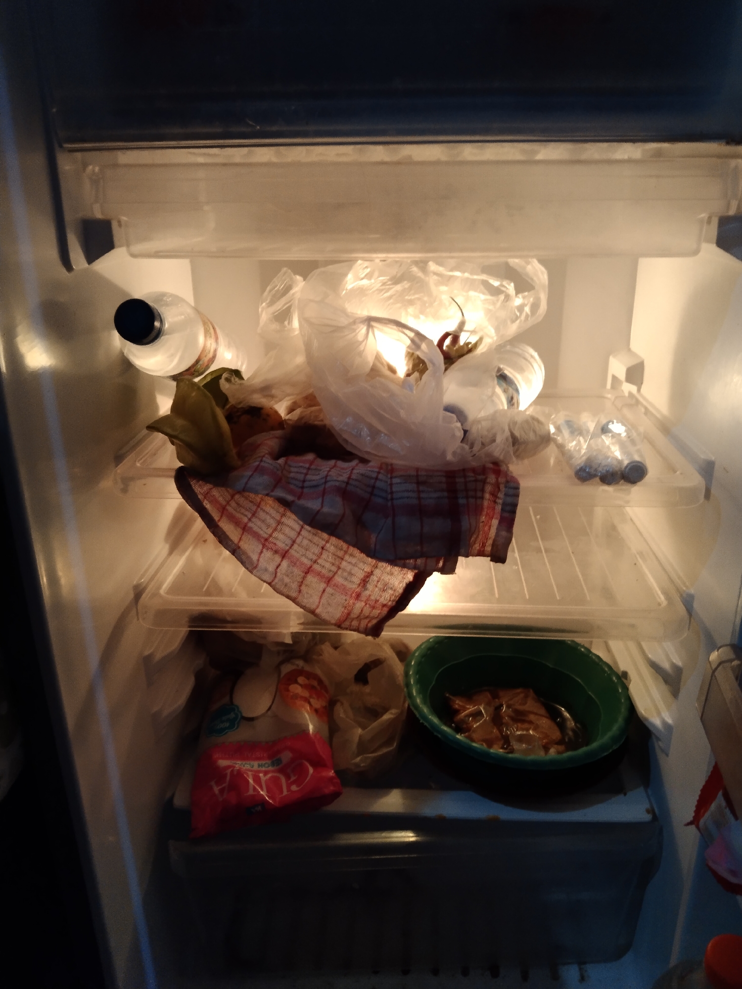 The image depicts the interior of a refrigerator, likely located in Buntalan, Indonesia. The refrigerator is moderately stocked with various food items and containers.
On the top shelf, there are several plastic bags, some of which appear to contain produce or other food items. A bottle with a black cap is visible on the left side of this shelf, along with a red and white checkered cloth draped over some items. A few smaller plastic bottles are arranged on the right side of the top shelf.
The second shelf from the top is mostly empty, with the checkered cloth extending onto it.
On the bottom shelf, a red plastic bag with white lettering is prominent on the left. To the right of this bag is a green bowl containing food items that appear to be submerged in liquid. The overall impression is one of a functional, albeit somewhat cluttered, refrigerator.