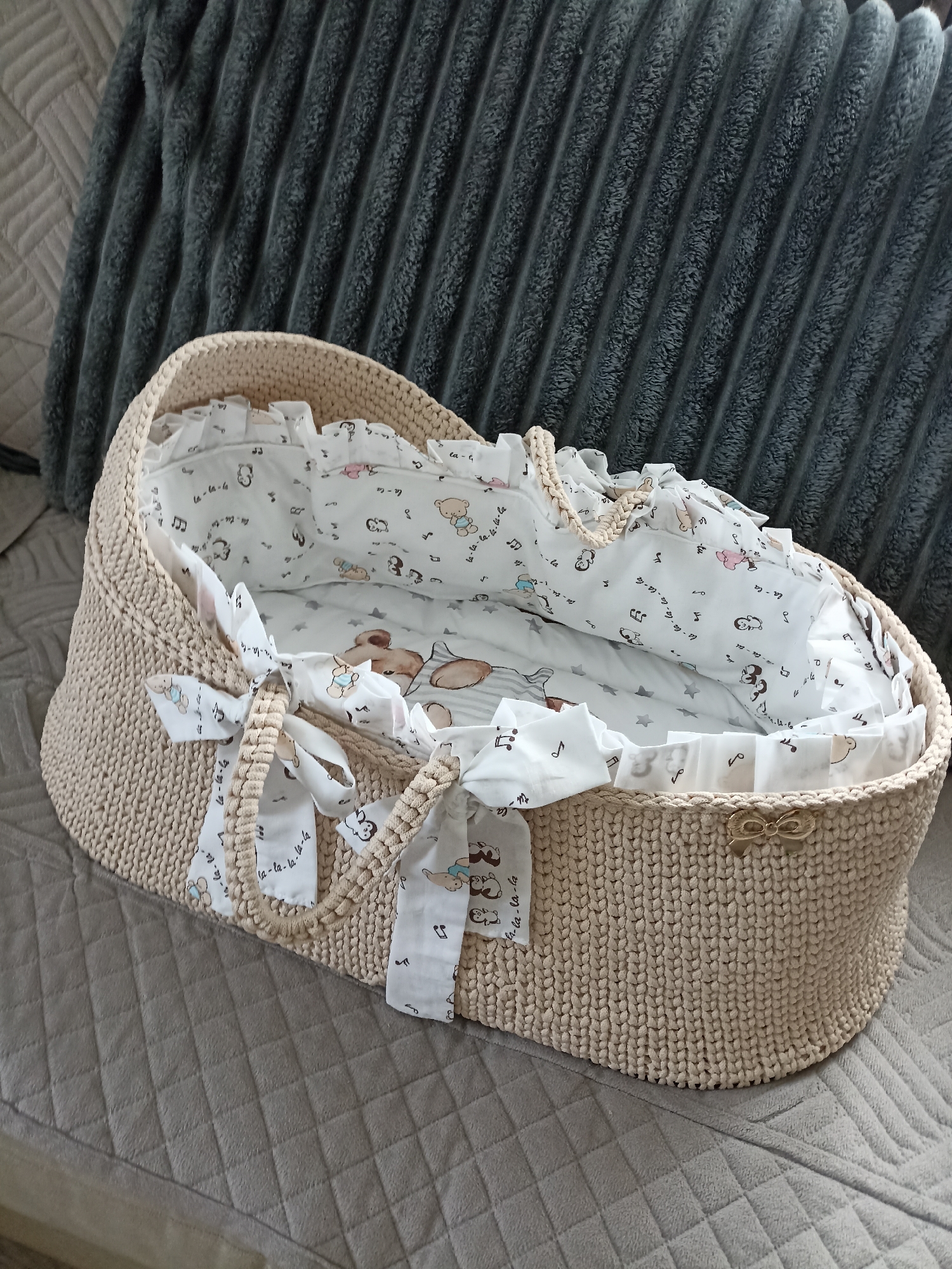 A crocheted bassinet with a beige exterior and a white fabric interior patterned with bears, musical notes, and stars rests on a quilted grey blanket. The bassinet has handles with fabric ties and a small gold bow on the outside. A dark grey, ribbed blanket is visible behind the bassinet, and the overall setting suggests an indoor environment, possibly a bedroom or living room. No people are present in the image.