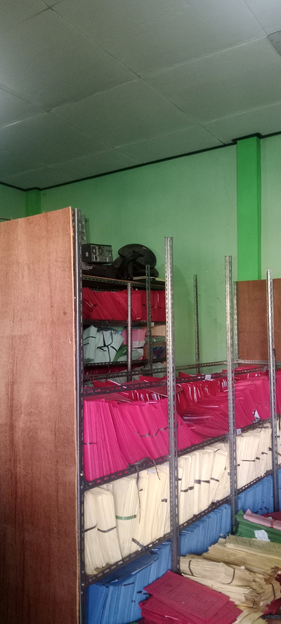 The image depicts a storage room with shelves filled with files. The shelves are made of metal, and the room has light green walls and a white ceiling with visible tiles and fluorescent lighting. On the top shelf, there are some electronic devices, including a radio or receiver, and a fan. The shelves are organized with various colored files: bright pink, light blue, yellow, and some green and red files are also visible. Some files are neatly stacked, while others appear to be loosely placed. A large wooden panel is on the left side of the image, partially obscuring the shelves. The overall impression is that of a disorganized yet functional archive or storage area. The context of Tuban, Indonesia, suggests this could be a municipal archive, a school's administrative storage, or a business's record-keeping facility.