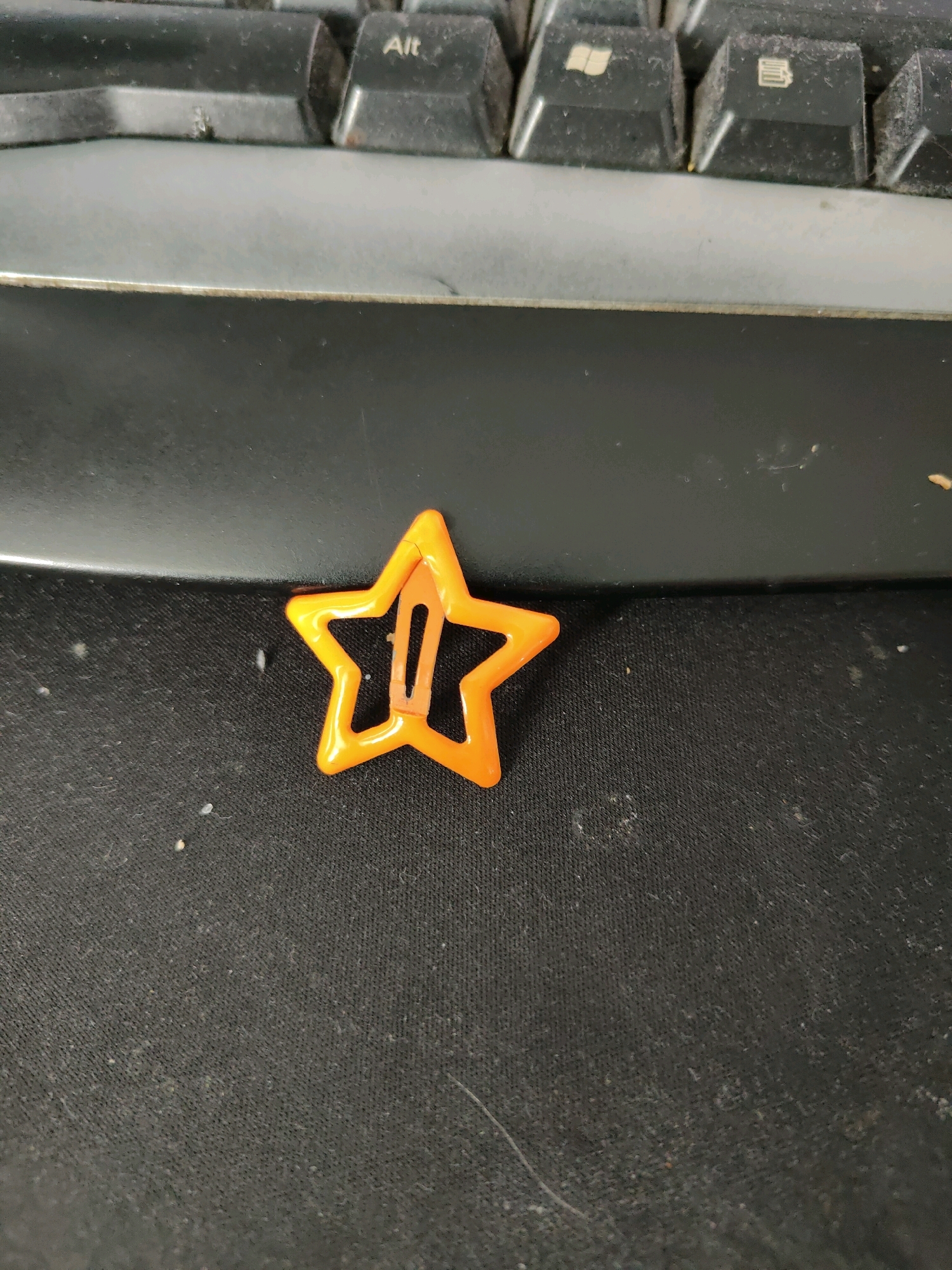 A single, orange, star-shaped hair clip is positioned in the center of the frame. The clip rests on a dark, textured surface, likely a fabric or mat, which is covered in a light dusting of debris. In the background, the edge of a black keyboard is visible. The lighting appears to be overhead, casting a slight shadow beneath the hair clip. The image is a close-up shot, with no discernible elements that would indicate a specific location within the city of Homyel, Belarus.
