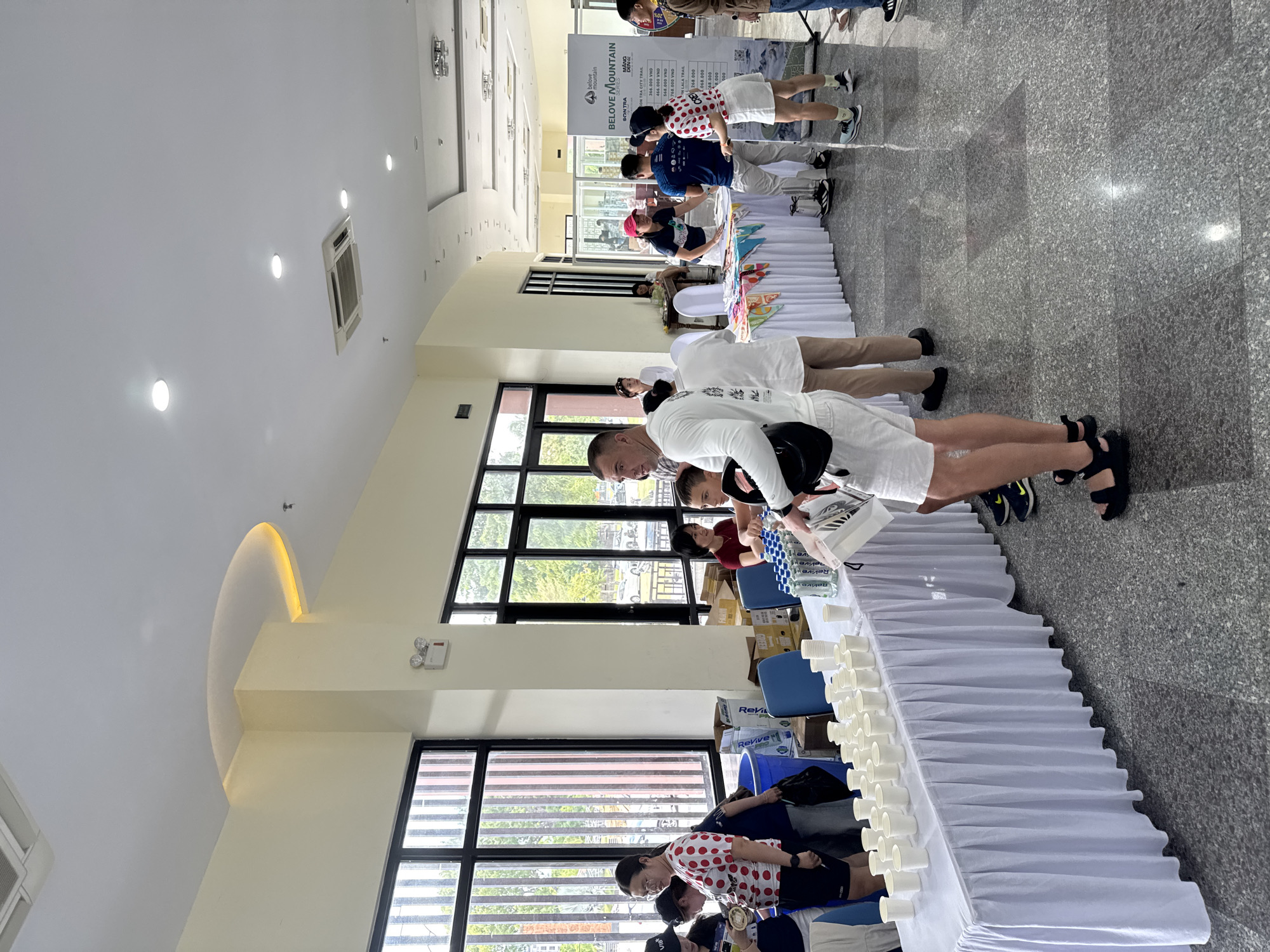 This image depicts an indoor event or registration area, likely associated with a race or outdoor activity, given the presence of a banner that reads "BELOVE MOUNTAIN SERIES" with the location "SON TRA". The scene is brightly lit with recessed ceiling lights and natural light filtering through large windows. The floor is a polished, speckled gray terrazzo.
In the foreground, a white table is draped with a white tablecloth and covered with rows of white paper cups, suggesting a refreshment station. To the right of this table, a man in white shorts and a white long-sleeved shirt is visible, appearing to interact with a white container. Behind him, another person in white shorts and black sandals is standing.
Further back, several people are gathered around other tables. One table has colorful patterned fabric draped over it, and a woman in a red shirt is interacting with attendees. A large banner displaying event information is positioned on the right side of the image. The overall atmosphere suggests an organized event with participants or staff attending to logistics and providing refreshments.
The location in the city of Hoi An, Vietnam, is not directly evident from the visual information within the image. However, the presence of the "SON TRA" location on the banner may refer to Son Tra Peninsula, a scenic area near Da Nang, which is a popular destination and often associated with events held in the wider region that includes Hoi An.