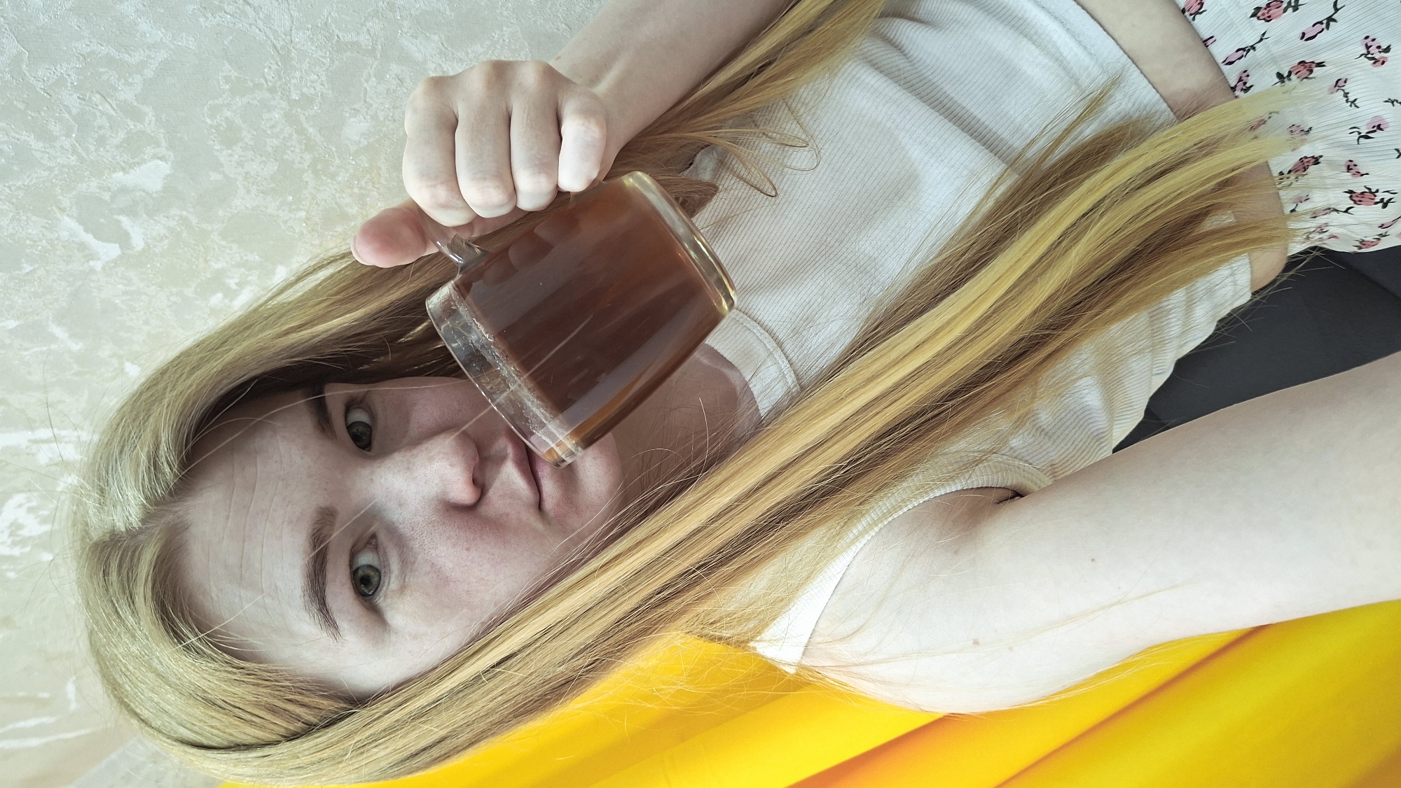 A fair-skinned woman with long blonde hair is holding a clear glass mug filled with a dark liquid, possibly coffee or tea. She is lying on what appears to be a yellow surface, with a floral patterned skirt visible on the right side of the frame. She is wearing a white top. The background is a textured, light-colored wall. Based on information available, it is not possible to determine the exact location within the city of Chaikovsky, Russia.