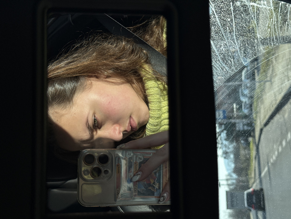 The image displays a car's interior rearview mirror reflecting a young woman taking a selfie, with an exterior view through the windshield below. The location is in the city of Banichan, Bulgaria.
The primary subject is a young woman, visible from the chest up in the mirror's reflection. She has brown hair, partially pulled back, and is wearing a bright, lime-green knitted turtleneck sweater and a black seatbelt across her chest. Her face, illuminated by strong sunlight from the right, exhibits a serious or contemplative expression with slightly furrowed brows. She is holding a clear-cased smartphone, resembling an iPhone, in her left hand, aimed towards the mirror. Two rectangular cards are visible within the phone's clear case. Her left hand shows manicured fingernails with a light base and darker, possibly metallic, designs.
Below the mirror, the car's windshield offers a view of the exterior environment. The windshield glass is densely covered with small, reflective water droplets or streaks. A paved road with a white dashed center line extends into the distance. A dark sedan is observed driving away on the left lane. On the right side of the road, parked vehicles are visible further down, along with leafless trees, utility poles, and a partially visible blue road sign. Distant, hazy mountains or hills are discernible on the horizon. The ambient lighting suggests a clear, sunny day.