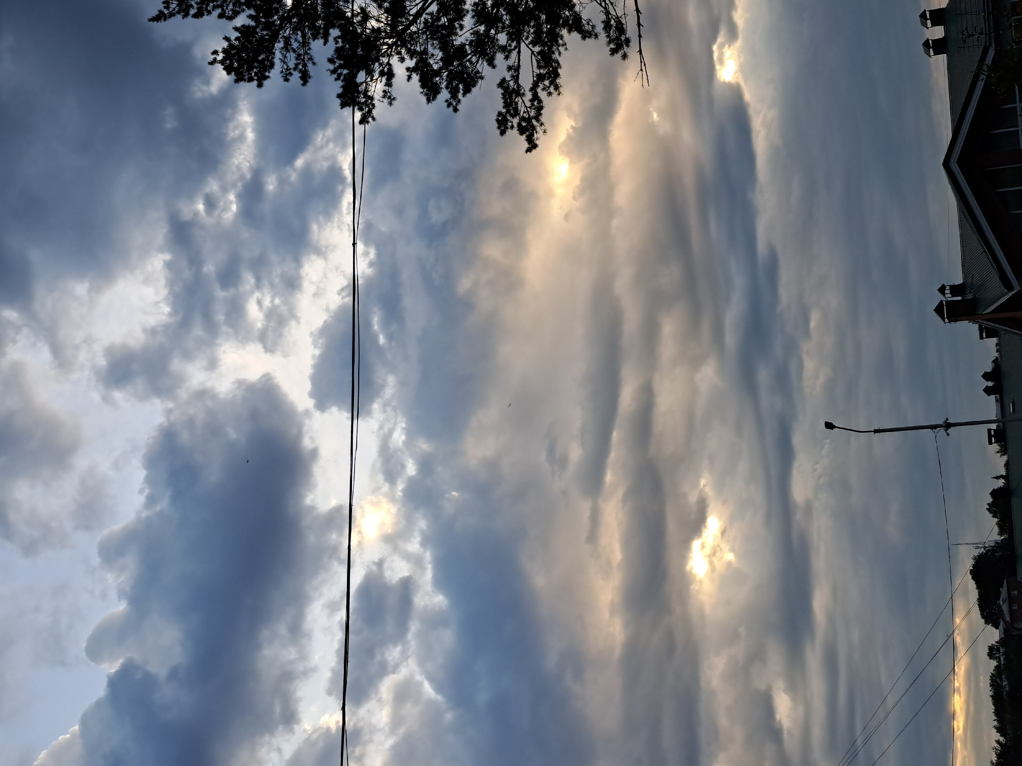 The image depicts a cloudy sky over a residential area, likely in Irkutsk, Russia, given the context. The sky is filled with heavy, dark gray clouds, with patches of sunlight breaking through in several areas, creating a dramatic effect. A silhouette of a coniferous tree is visible on the right side of the frame, its branches extending towards the sky. In the foreground, the roofs of houses, characterized by dark, sloped surfaces and visible chimneys with caps, are silhouetted against the sky. Utility lines stretch horizontally across the upper portion of the image. A street lamp and what appears to be a surveillance camera are mounted on a pole in the lower middle section of the frame. The overall impression is of a dramatic sky at dusk or during an overcast day, common in temperate climates.
