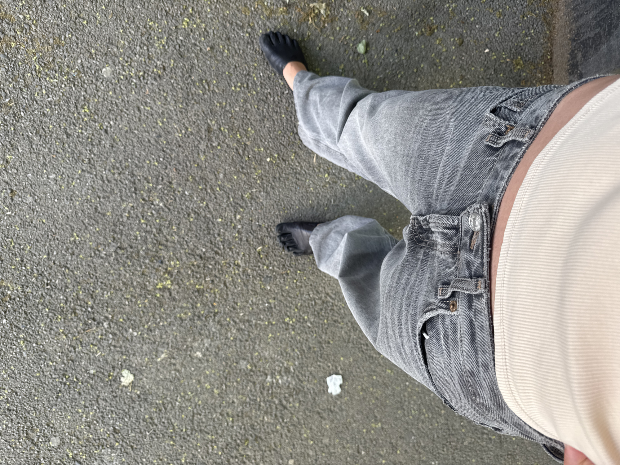 An overhead shot captures the lower portion of a person wearing grey, wide-legged jeans, a light-colored, horizontally ribbed top, and black tabi shoes, standing on paved ground with scattered debris. The location cannot be determined.
