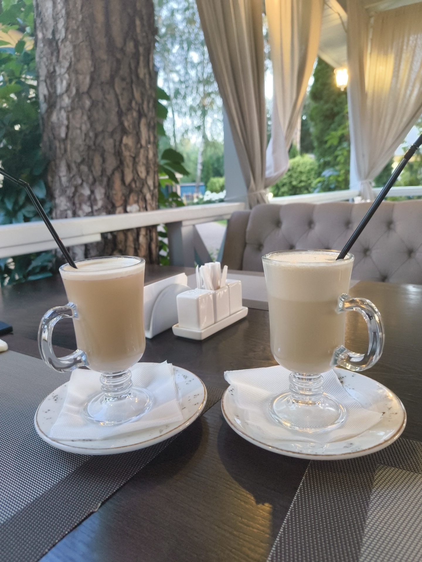 The image shows two glasses of latte on a dark wooden table in an outdoor cafe in Ramenskoye, Russia. The glasses are tall with handles and contain a layered coffee drink topped with foam. Black straws are present in each glass. The glasses are placed on white saucers with decorative speckles, each with a folded white napkin underneath. In the background, there is a quilted upholstered bench seat and sheer curtains hanging from a canopy, suggesting an outdoor seating area. A tree trunk is visible on the left side of the frame. A white napkin dispenser with napkins and two white condiment shakers are on the table between the two drinks. The lighting appears to be natural, indicating it is daytime.