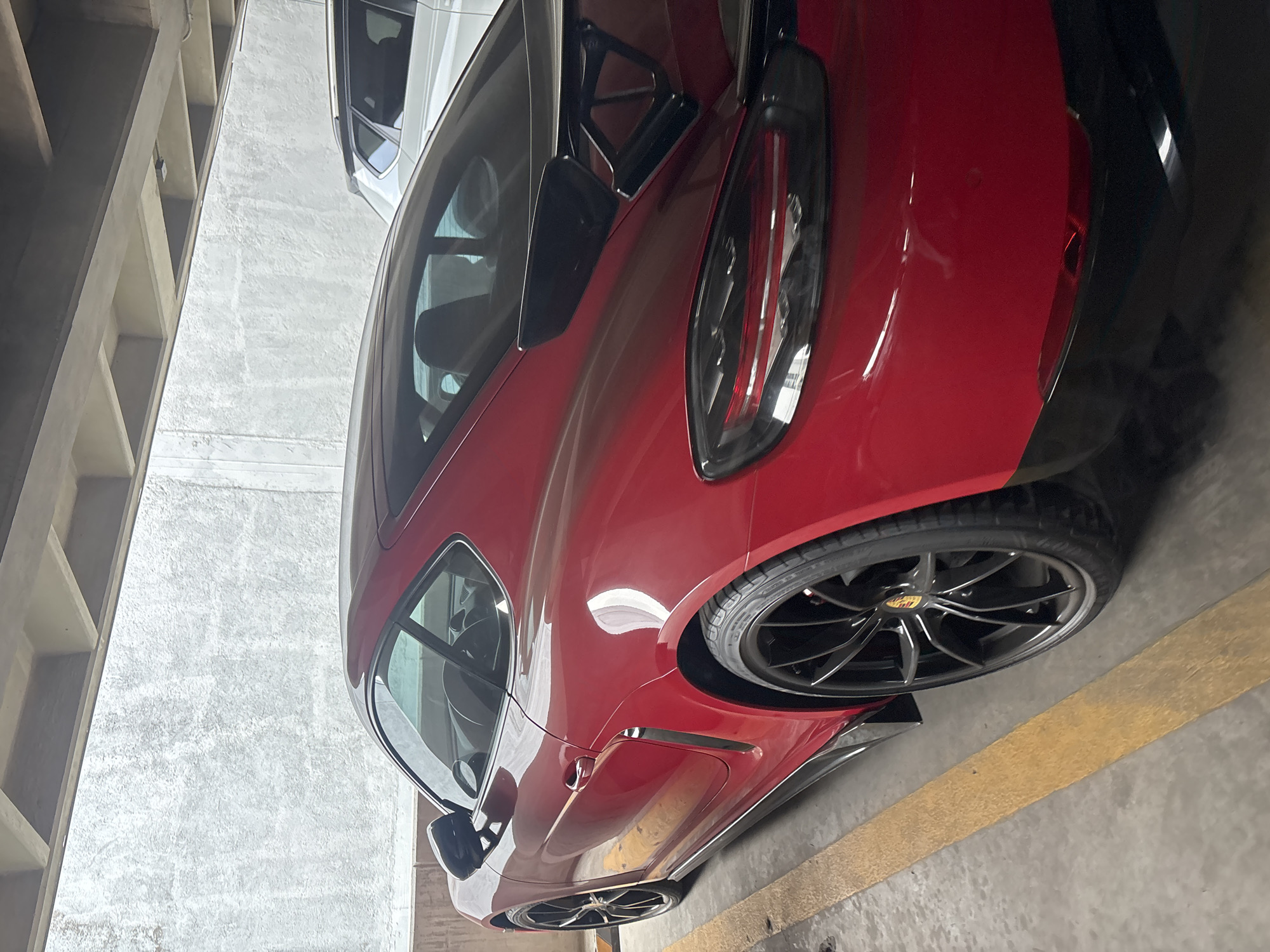 The image shows a red Porsche parked in a parking garage. The car is parked between a white car and a concrete wall. The parking garage is located in San Bernardino Tlaxcalancingo, Mexico. The image captures the car's sleek design, including its rear lights and wheels. The yellow parking lines indicate designated parking spaces. No people are visible in the image.