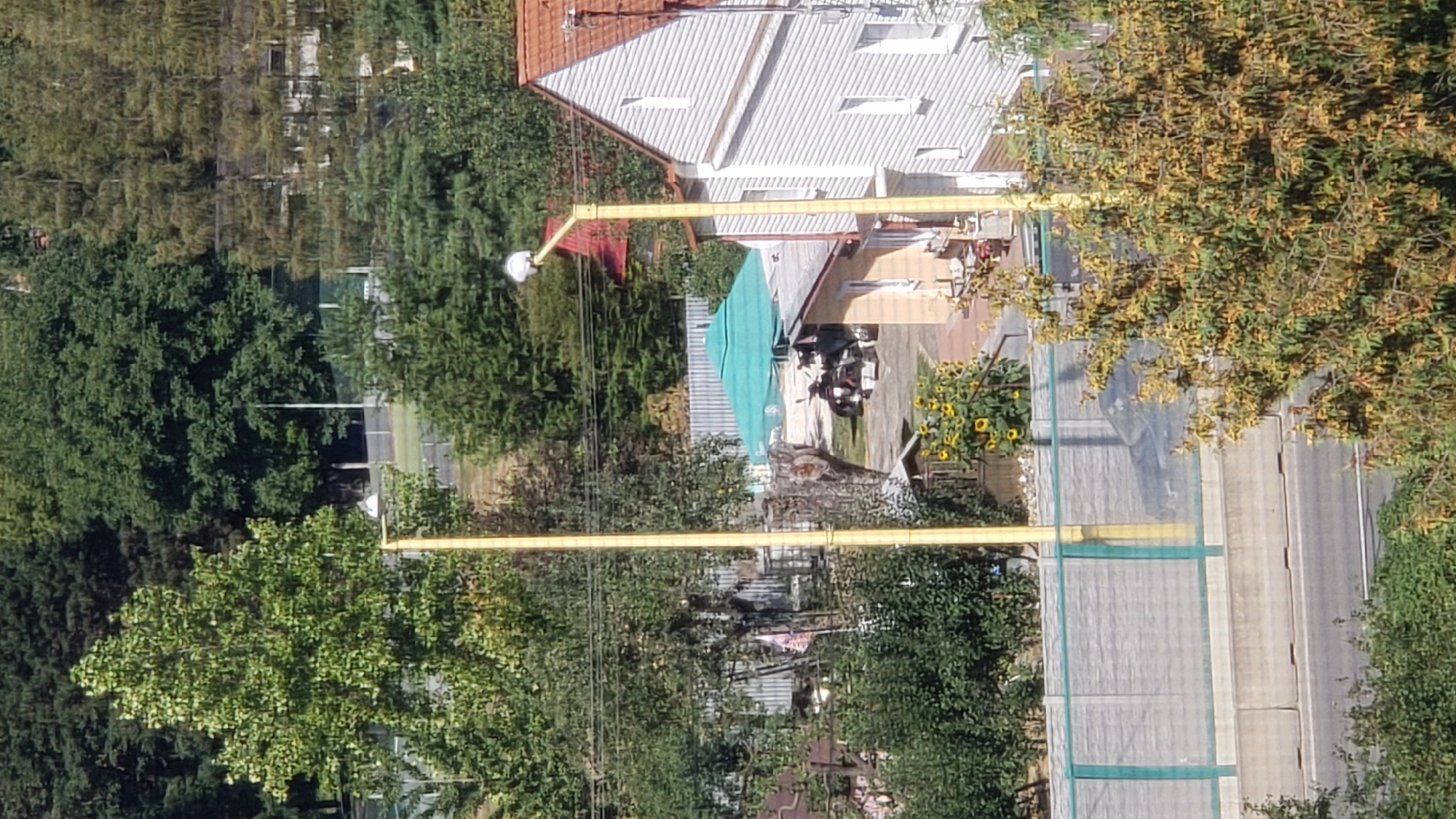 This is an outdoor, eye-level, daytime, medium shot photograph taken from a slightly elevated position. The scene depicts a residential area with elements of nature and urban infrastructure. In the foreground, a paved road runs horizontally across the bottom of the frame, bordered by a concrete barrier and green fencing. Beyond the fencing, a large tree with yellowing leaves occupies the right side of the image.
Behind the barrier, there's a light-colored concrete wall with a green net overlay. A tall, yellow lamppost stands near the center of the frame, its top featuring a white, spherical light fixture. To the left of the lamppost, a large, leafy green tree is prominent, with a flag visible amongst its branches. Further into the midground, a light-colored residential building with a red-tiled roof and white siding is partially visible on the right.
In the center of the midground, a large green umbrella provides shade over an outdoor seating area. Underneath the umbrella, a motorcycle and what appears to be a scooter are parked. A smaller, single-story building with a light-colored facade is situated to the left of the umbrella. A cluster of sunflowers is visible to the left of this building.
The background is dominated by dense, green trees, indicating a wooded or park-like environment. The overall atmosphere is that of a sunny, possibly autumn day. The location is identified as being in Prešov, Slovakia, suggesting a European urban or suburban setting. There are no people clearly visible in the scene.