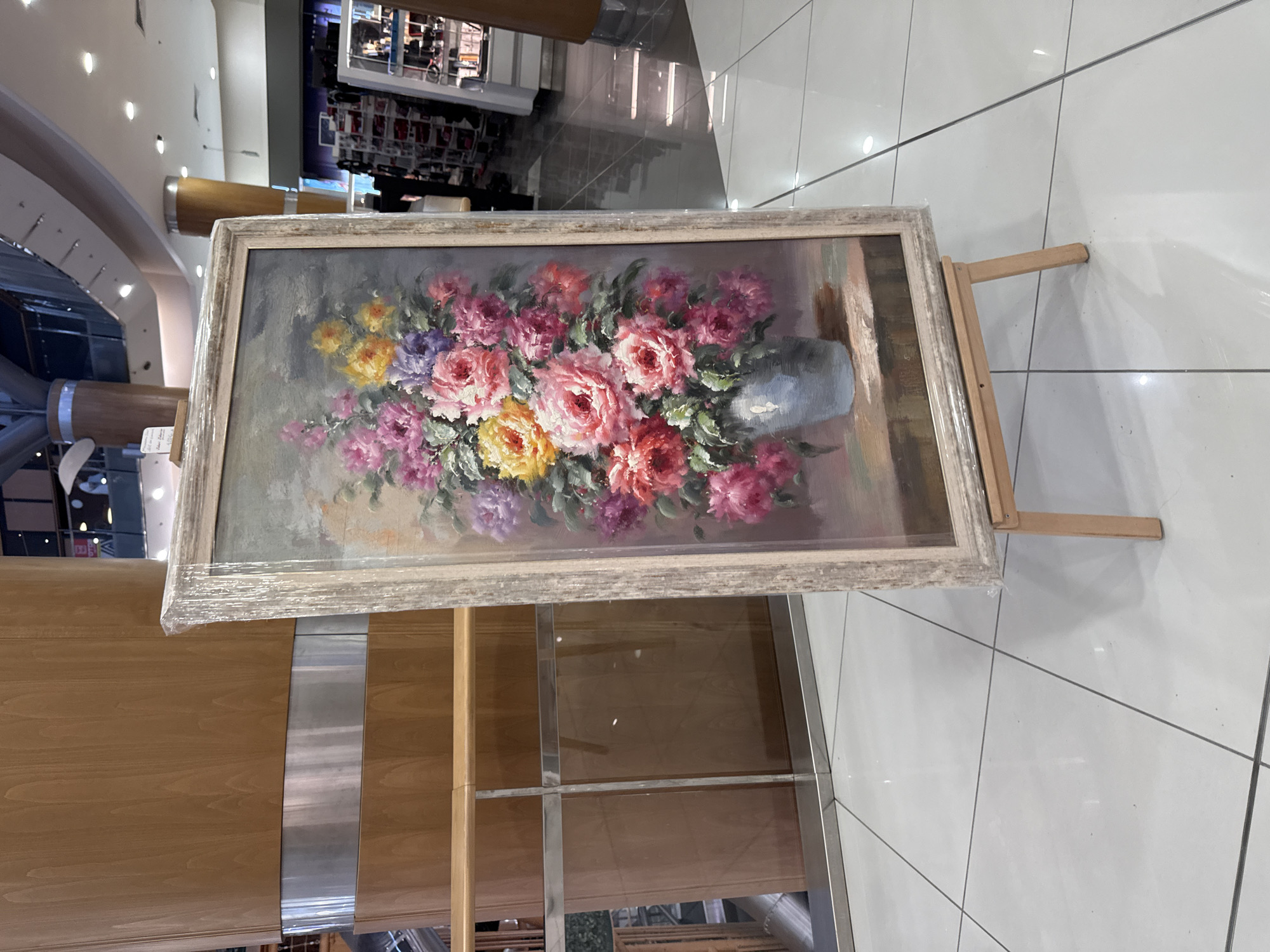 The image shows a framed oil painting of a floral arrangement on an easel, placed on a tiled floor in what appears to be a public indoor space, possibly a shopping mall or a gallery. The painting depicts a vase filled with various colorful flowers, including pink, purple, yellow, and red roses. The easel is made of light-colored wood and is positioned in front of a wooden pillar with a metallic band. In the background, there are escalators, railings, and display shelves, suggesting a commercial or public environment. The painting is partially covered with plastic wrap.