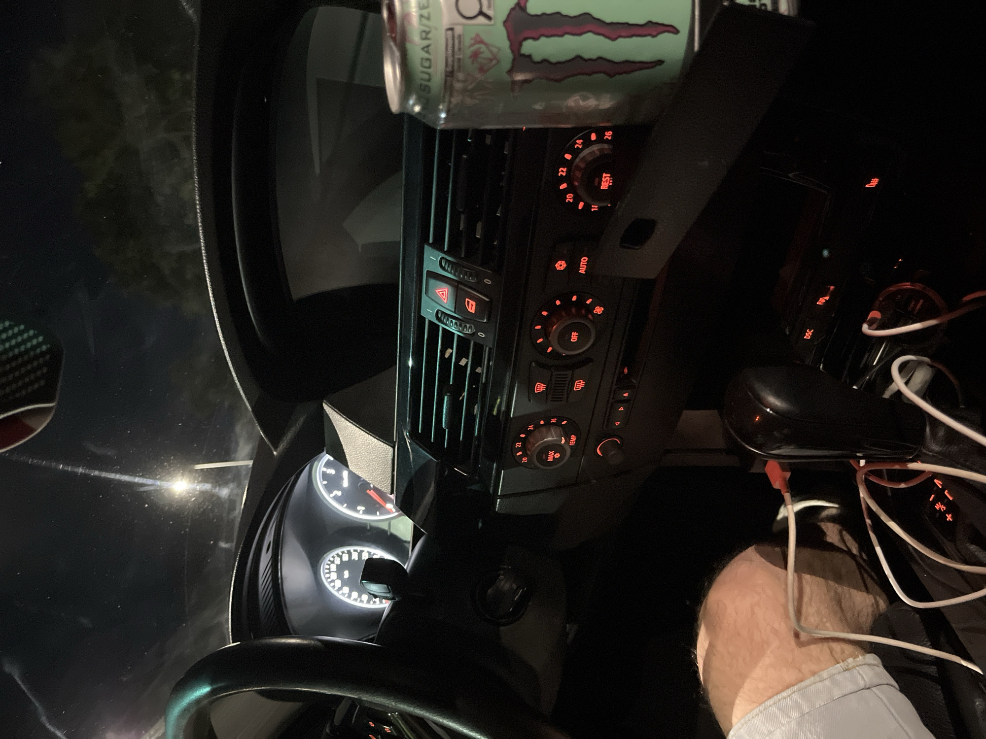 The image shows the interior of a car. The dashboard features climate control knobs and buttons illuminated in red. A can of Monster Energy Sugar/Zero drink sits on top of the central air vents. The instrument cluster, including the speedometer and tachometer, is visible behind the steering wheel. To the right, a gear shift and assorted cables are present, along with a person's knee and part of their leg clad in a light-colored garment. The location, Shumen, Bulgaria, cannot be confirmed from the visual information alone.
