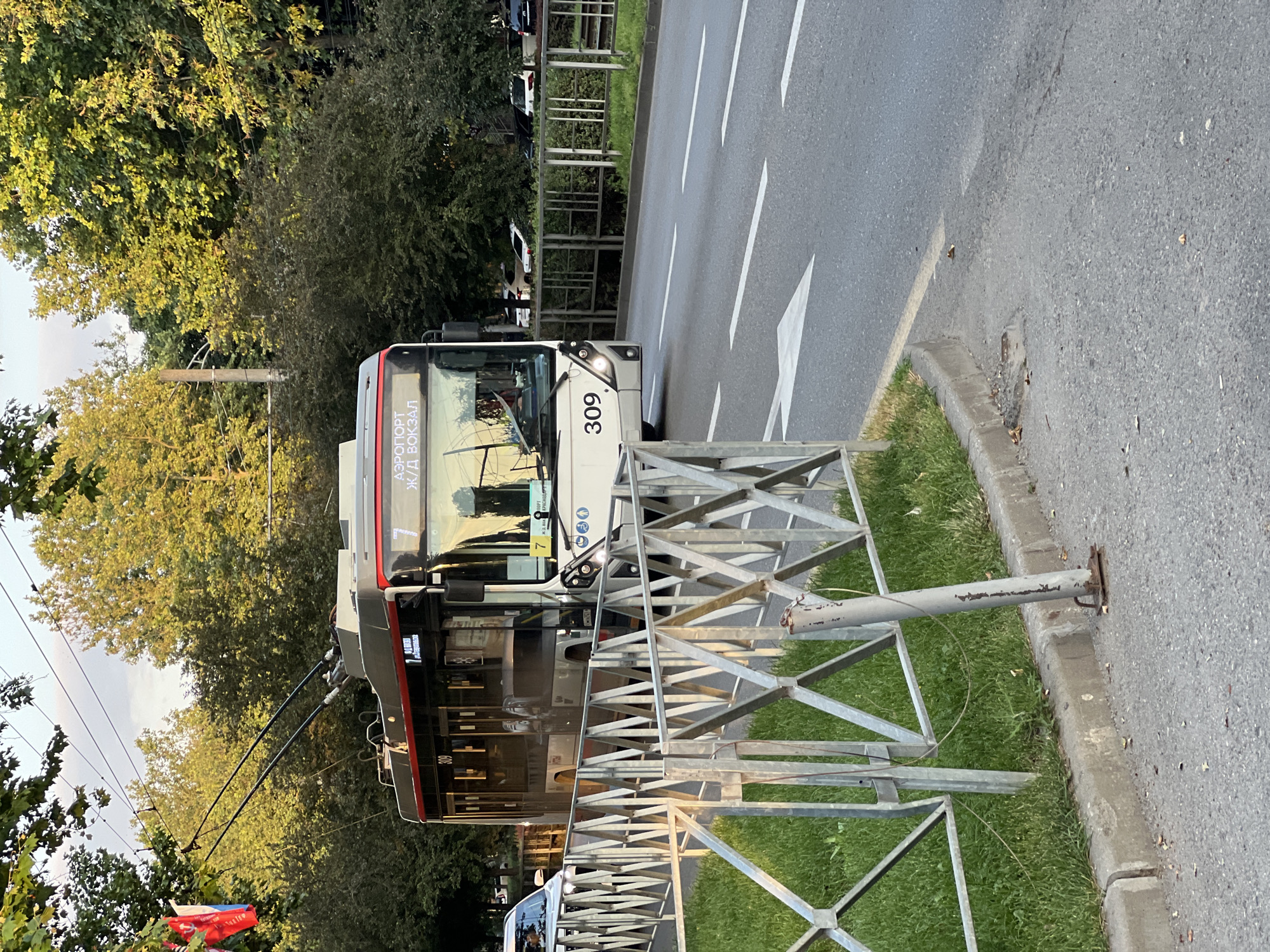 A trolleybus with the number 309 is driving on a city street. The trolleybus is black and white with a red stripe. The destination displayed on the front of the trolleybus is "АЭРОПОРТ Ж/Д ВОКЗАЛ" which translates to "Airport Railway Station". The trolleybus is powered by overhead electrical wires. In the background, there are trees and buildings. In the foreground, there is a metal fence and a sidewalk. A white car is parked on the side of the road. There are traffic signs and road markings on the street. The image appears to be taken during the daytime.