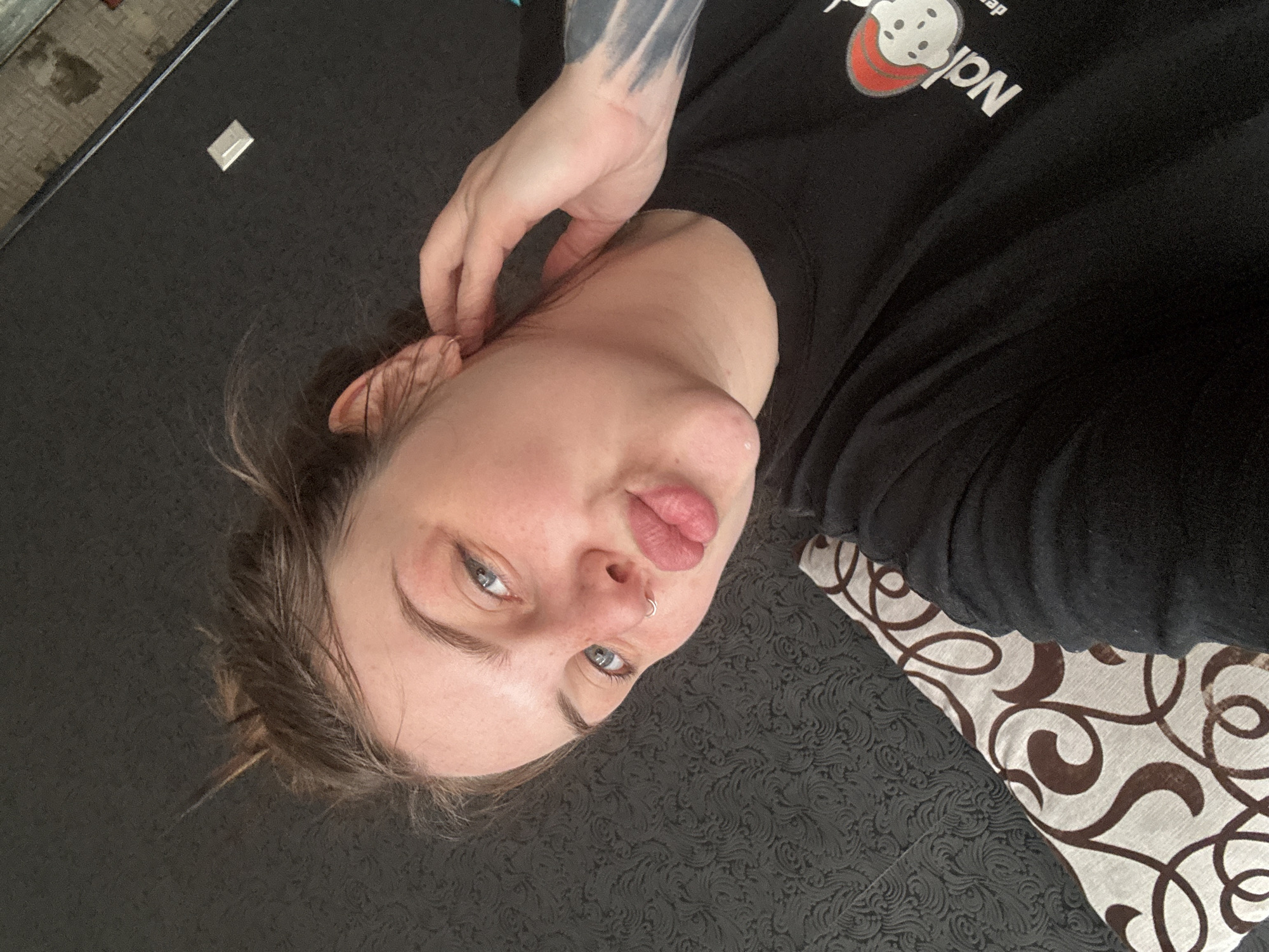 A young person with light skin and blue eyes lies on a textured surface, partially covered by a patterned pillow or fabric. The person has a nose ring and is wearing a black t-shirt with white and red lettering and a logo. The individual's left arm, featuring a dark tattoo, is raised near their head. A small rectangular object is visible on the textured surface near the person's head. The image is taken from a slightly overhead, angled perspective. There is no information available to indicate the location is Chornomorskoe, Ukraine.