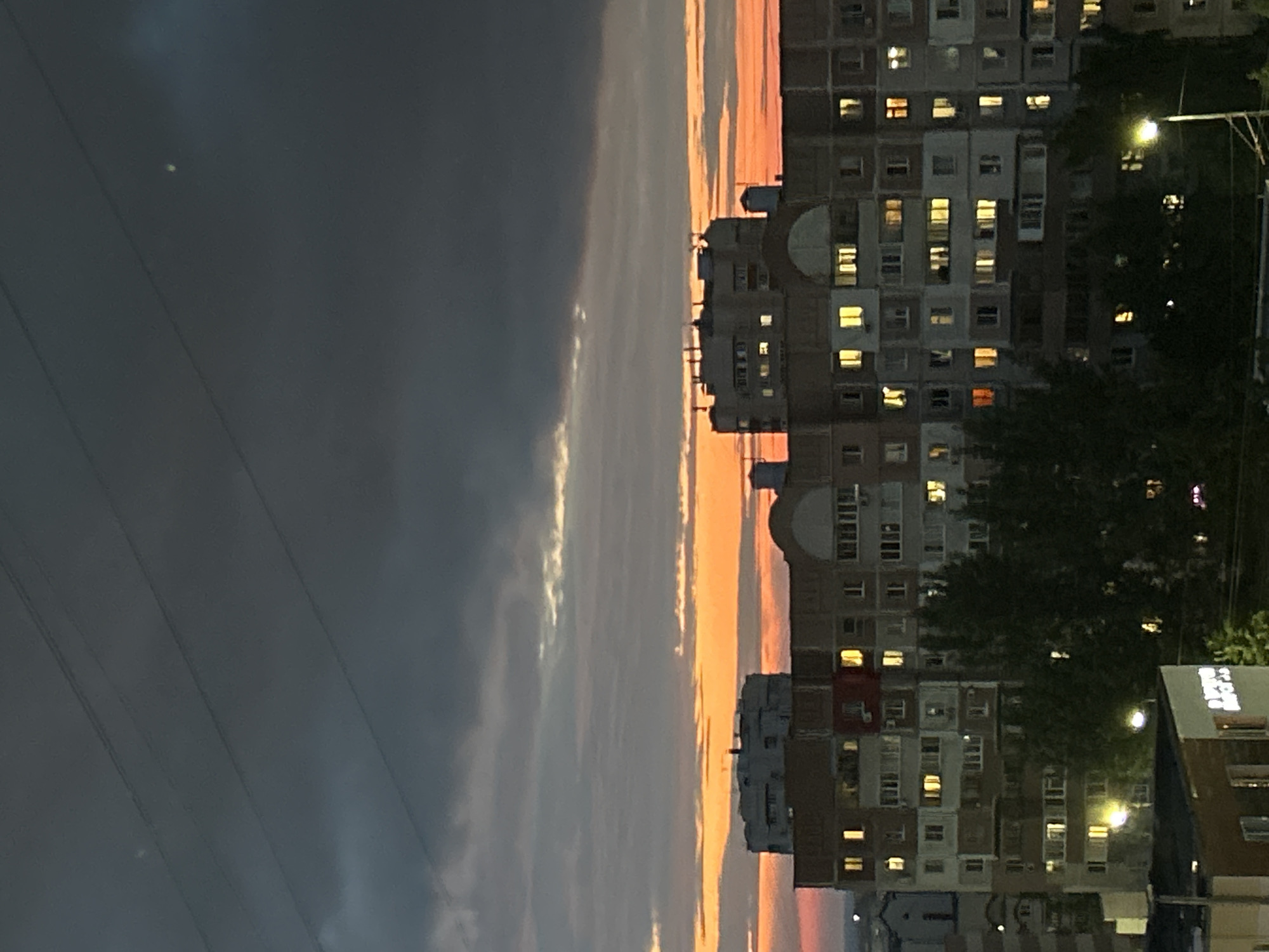 This is a medium shot, eye-level photograph taken at dusk in an urban environment. The foreground is dominated by the dark silhouettes of several buildings, with trees obscuring portions of their lower sections. The buildings are multi-story residential structures with numerous windows, some of which are illuminated with warm, yellow light, suggesting occupancy. A street lamp is visible on the right side of the image, casting a bright light.
The midground features more residential buildings, also appearing to be multi-story apartment complexes. These buildings are also partially obscured by trees, and their windows are a mix of lit and unlit. The architecture is typical of Soviet-era or post-Soviet era mass housing.
The background is characterized by a dramatic sky. The upper portion of the sky is a deep, dark gray with hints of charcoal, possibly indicating storm clouds or heavy cloud cover. Below this, a vibrant band of orange and yellow stretches across the horizon, a characteristic of a sunset or sunrise. The color gradient suggests the sun is low on the horizon, casting its last rays. Several thin, dark lines, possibly power lines, are visible stretching diagonally across the dark upper portion of the sky. The overall mood is somewhat somber yet also visually striking due to the contrast between the dark sky, the glowing windows, and the vibrant sunset colors.