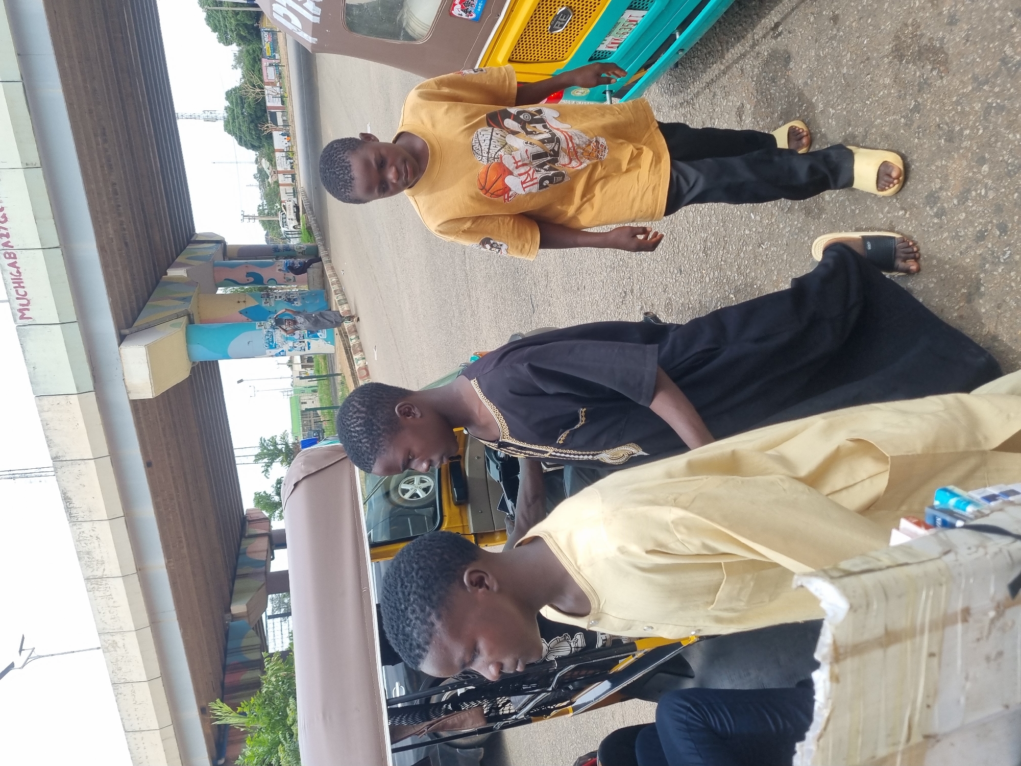 Three young men are standing near a yellow, blue, and beige commercial tricycle (keke) on a paved street in Jos, Nigeria. An overpass structure with graffiti on the pillars is visible in the background. The first young man is wearing a yellow t-shirt with a basketball design and black pants with yellow sandals. The second young man is wearing a black traditional shirt with gold trim and black pants with black sandals. The third young man is wearing a light yellow traditional shirt and blue jeans. The first young man is standing facing the camera. The other two young men are interacting with the tricycle.