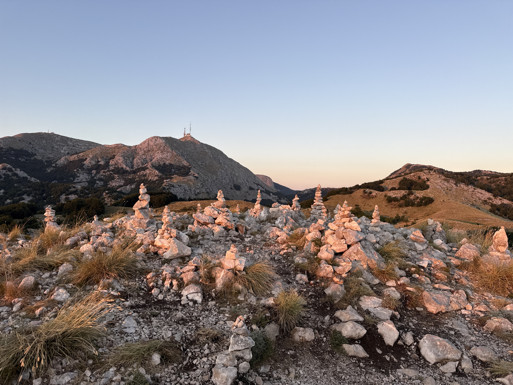 The image depicts a rocky mountainous landscape at sunset. In the foreground, numerous cairns, or stacked stone piles, are scattered across the uneven terrain. These cairns vary in height and size, with some being quite tall and others small stacks of just a few stones. The ground is covered with smaller rocks, pebbles, and patches of dry, golden grass.
In the middle ground, more cairns are visible, leading the eye towards the background. The mountains in the distance are rugged and barren, with some vegetation visible in the valleys. One prominent mountain peak in the mid-background features a structure with antennas on its summit, suggesting it might be a telecommunications tower or observation point. The sky above is clear and a pale blue, transitioning to a soft orange hue near the horizon, characteristic of sunset. The overall scene is natural and appears to be a remote, elevated location.
The location is identified as Mount Lovćen, near the village of Njeguši, in the municipality of Cetinje, Montenegro. This area is known for its significant cultural and historical importance, being the birthplace of the Petrović-Njegoš dynasty, rulers of Montenegro. The cairns are likely trail markers or memorial markers placed by visitors. There are no people or animals visible in the image. The lighting suggests it is either sunrise or sunset, with the warm tones of the sunlight illuminating the rocks and vegetation.