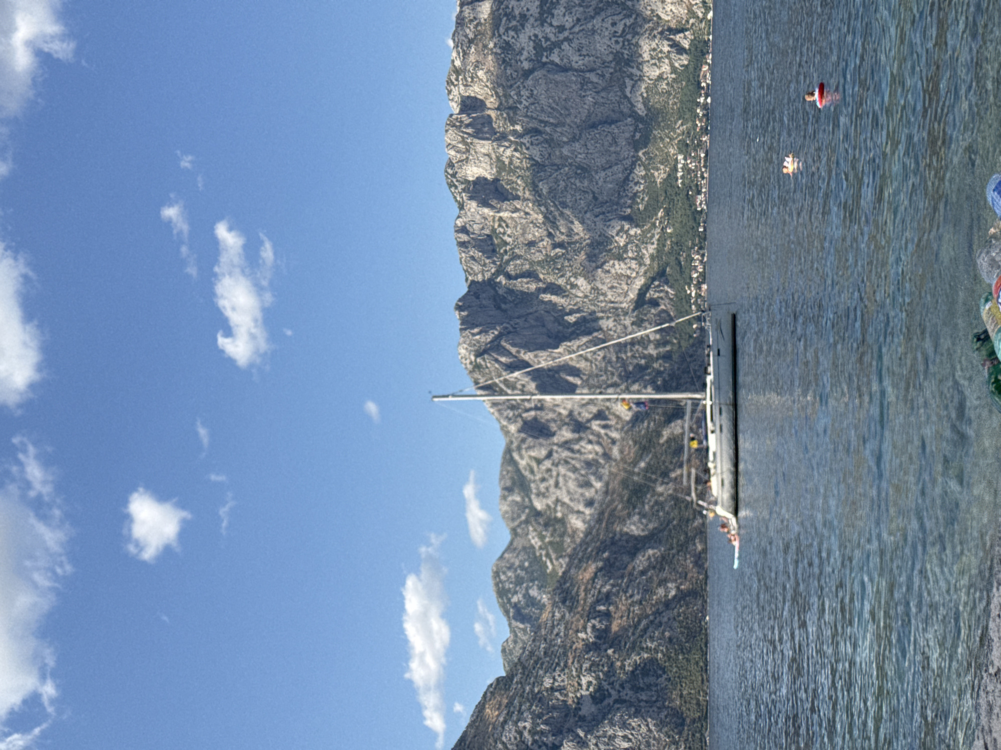 In the city of Prčanj, Montenegro, a clear, bright day unfolds with a sailboat gliding on the calm, blue waters of the bay. Towering, rocky mountains form a dramatic backdrop, their rugged slopes dotted with green vegetation and small settlements. A white sailboat, its mast reaching towards the sky, is anchored in the mid-ground. Several people are visible on the deck of the sailboat. Further out in the water, two individuals are swimming, one using a red inflatable ring. The foreground shows a rocky shoreline with some colorful objects, possibly beach accessories, scattered on it. The sky is a vivid blue with scattered white clouds.