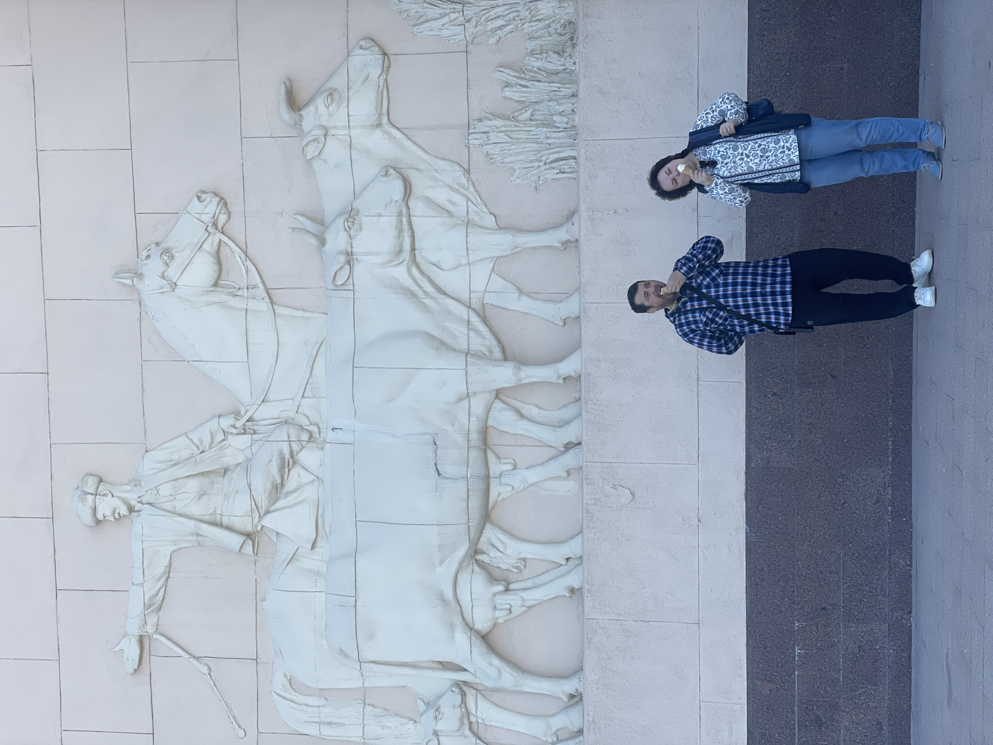 Two people are standing to the right of a large bas-relief on a light-colored wall. The bas-relief depicts a man on horseback, with two cows behind him. The man on the horse is wearing a long coat and hat, and he is holding a long stick. The cows are standing next to each other. The people are standing next to the wall. The man is wearing a blue plaid shirt and black pants. The woman is wearing a white shirt with a floral pattern, a black vest, and blue jeans. Both people are eating ice cream cones. A dark stone or concrete path runs along the right edge of the wall. The location appears to be in Moscow, Russia.