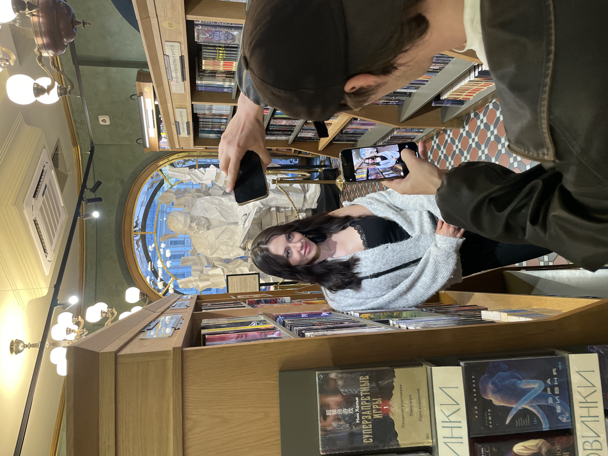 In Saint Petersburg, Russia, a young woman with long dark hair and a black lace-trimmed top, layered with a grey cardigan, poses in front of a bookshelf. She is in the "Dom Knigi" (House of Books) department store, a prominent landmark on Nevsky Prospekt. Two individuals are documenting the scene. One, from a first-person perspective, is holding a smartphone at eye level, capturing a photo of the woman. Another, also holding a smartphone, is taking a photograph of the same woman, framing her from the chest up against a backdrop of books and a large, white, abstract sculpture of a man. The interior of the store features wooden bookshelves, ornate chandeliers, and a patterned tiled floor.